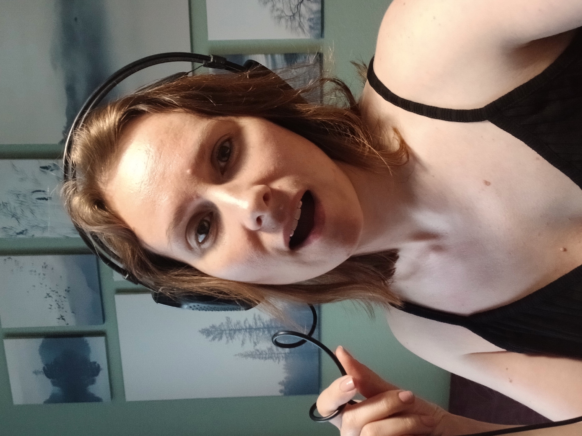 The image shows a close-up, selfie-style shot of a woman with light brown hair, wearing black headphones and a black tank top. She is holding a coiled black cable in her left hand, which is positioned near her mouth. Her mouth is open, and she appears to be speaking or singing. The background consists of a light green wall adorned with four framed abstract landscape artworks. The image was taken indoors, likely in a residential setting, and there is no discernible information to suggest a specific location within the city of Yekaterinburg, Russia.