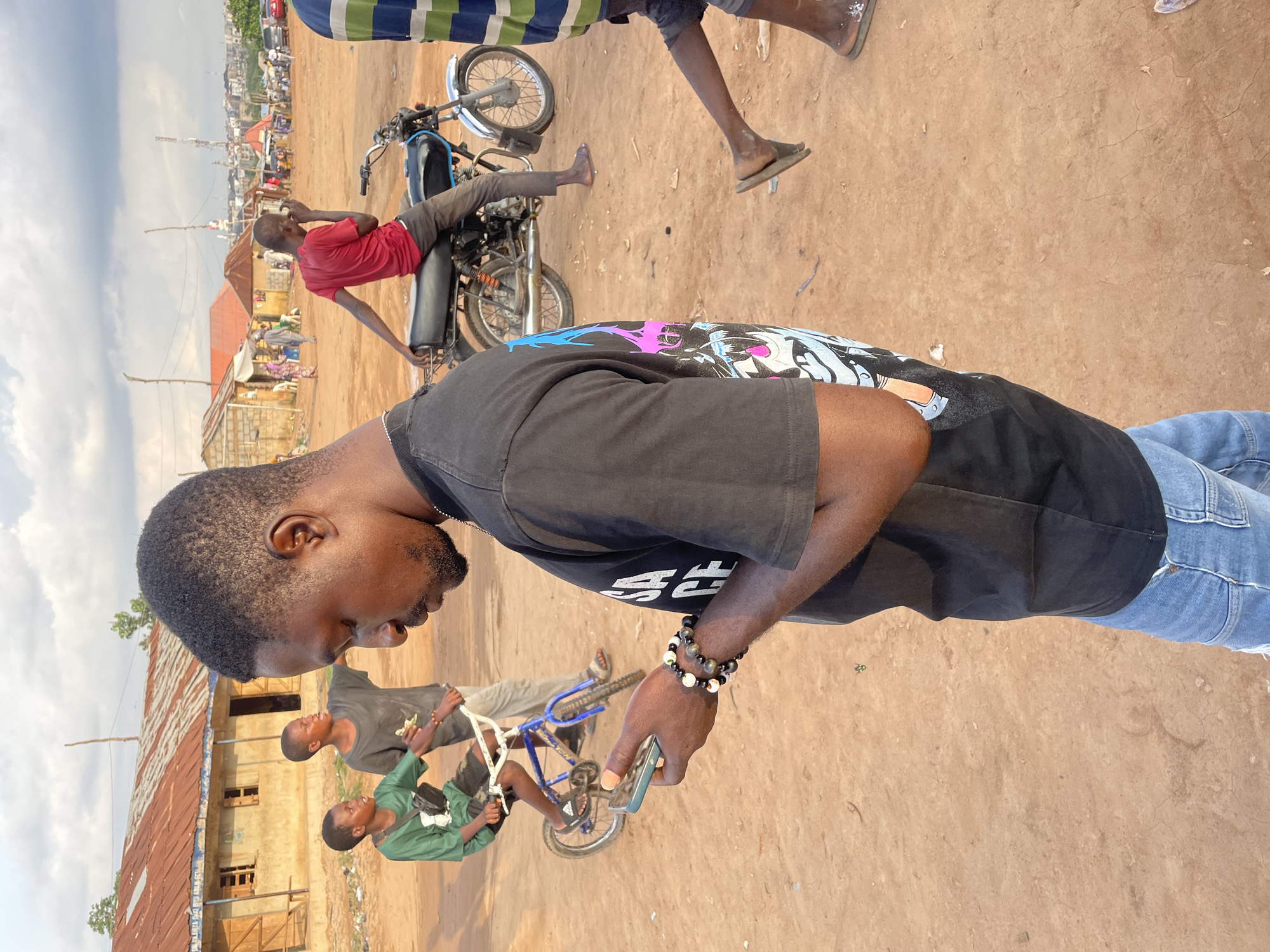 In Abuja, Nigeria, a group of young boys are seen playing and socializing in an unpaved, dusty area. One boy, wearing a green shirt, is on a small blue bicycle with white handlebars. Another boy, in a dark t-shirt, stands beside him. In the foreground, a young man in a black t-shirt and blue jeans is looking at his phone. He has bracelets on his wrist. In the background, a red motorcycle is parked, with a young man in a red t-shirt sitting on it. Further in the background, there are several buildings with corrugated metal roofs and a distant cityscape. The sky is partly cloudy.