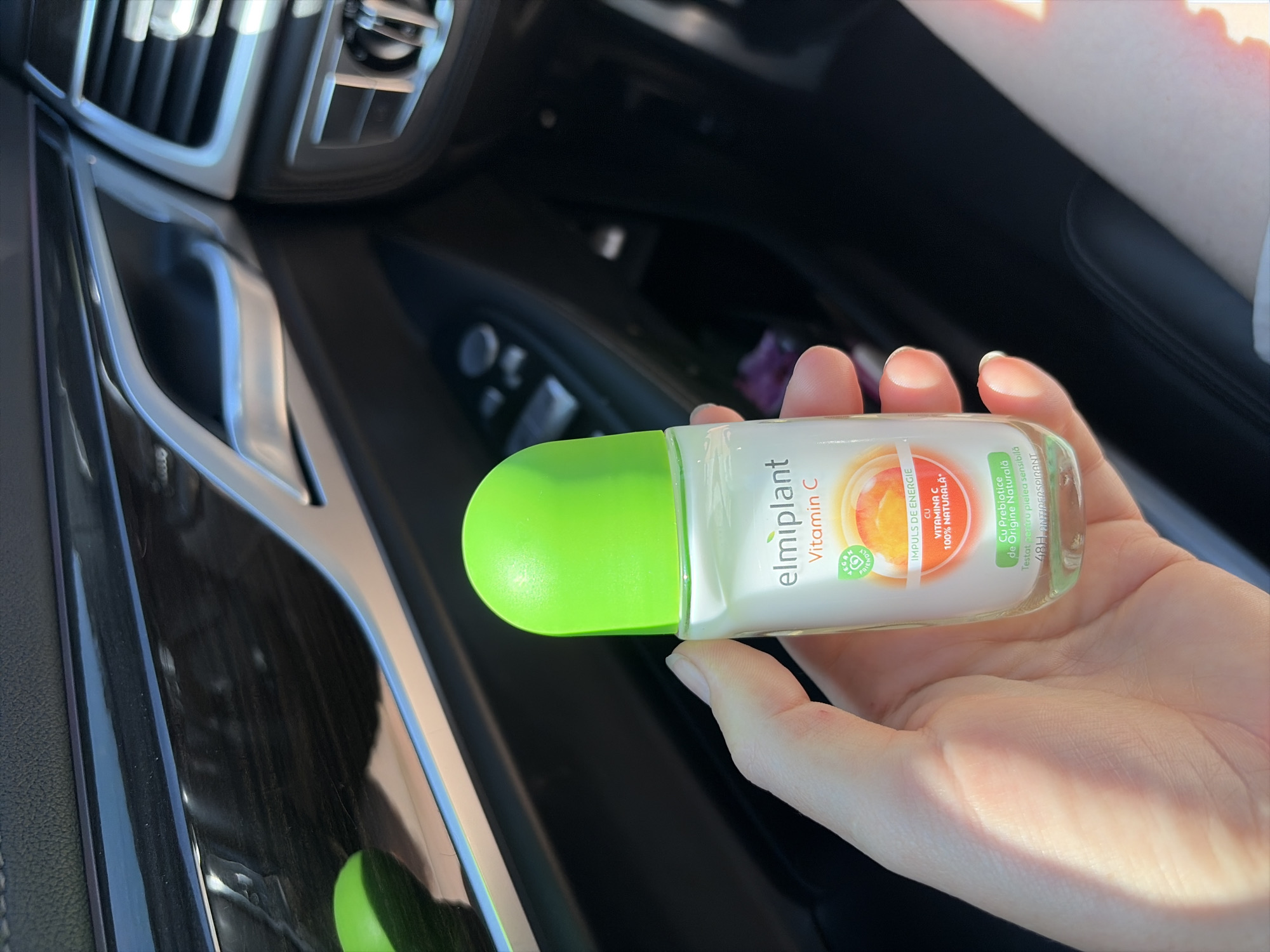 A person's hand holds a small, clear plastic bottle with a bright green cap and a white label featuring the brand "elmiplant" and "Vitamin C." The bottle is positioned in the foreground, against a blurred background of a car's interior. The background shows dark leather upholstery, a metallic trim, and a car's air vent. The scene suggests the bottle is an underarm deodorant given the text "48H ANTIPERSPIRANT" visible on the label. The overall lighting is bright, indicating daylight. The location is identified as being in Criuleni, Moldova, though this is not visually discernible from the image itself.