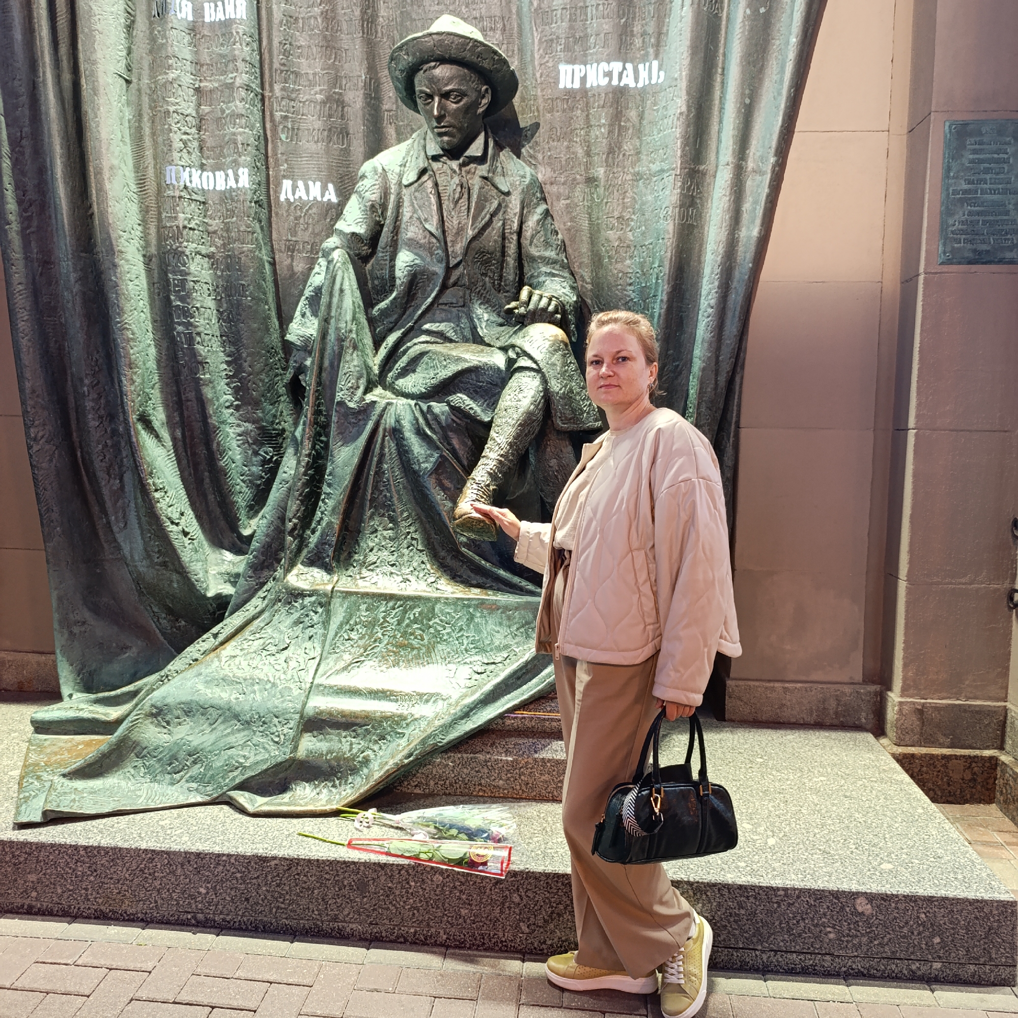 A woman stands to the right of a large bronze statue of a seated man wearing a hat and a suit. The statue is positioned in front of a backdrop with draped fabric imagery, which is inscribed with text. The woman is wearing a light beige quilted jacket, a beige t-shirt, and tan pants. She is holding a dark handbag and her right hand is touching the statue's leg. In front of the statue's base, on the ground, are two red roses in clear plastic wrapping. The scene appears to be an outdoor public space with a paved walkway in the foreground and a building to the right. The statue is likely a monument to a notable figure, given the inscriptions on the backdrop. Based on the inscriptions visible on the backdrop of the statue, such as "ПРИСТАНЬ" (Pier/Haven) and other text in Russian, this statue is likely located in Moscow, Russia, and is a monument to the writer Ivan Bunin, who is often associated with themes of exile and longing for a homeland, evoked by the "Pier/Haven" metaphor.
