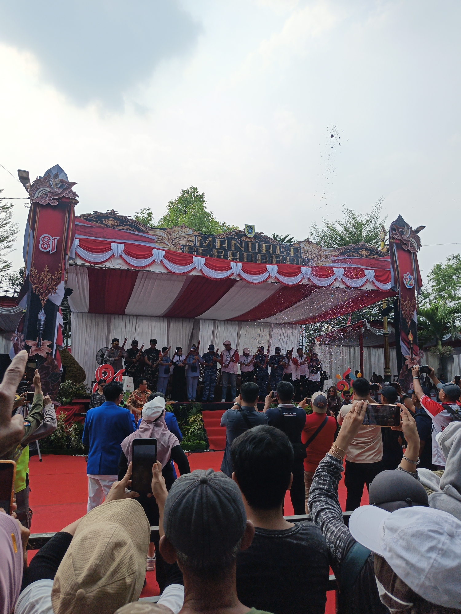 A group of people are gathered to watch a performance on a stage decorated with red and white banners. Some individuals in the crowd are taking pictures with their phones, while others are watching the event. Confetti is falling from above the stage, suggesting a celebratory atmosphere. The backdrop includes trees and a cloudy sky. The event appears to be taking place in Klaten, Indonesia, as indicated by the presence of banners with the number "81" and other Indonesian cultural motifs.