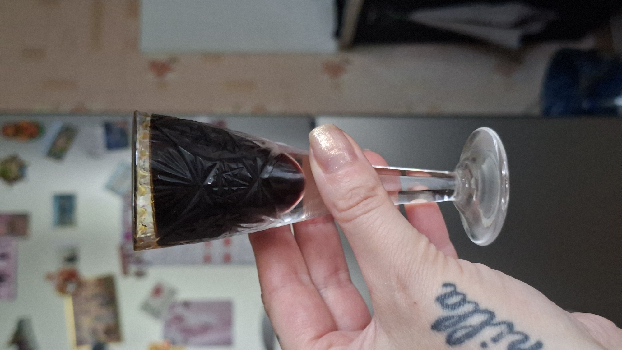A close-up, eye-level shot captures a person's hand holding a small, clear glass filled with a dark liquid, possibly liqueur or coffee, against a blurry background of a kitchen refrigerator adorned with various magnets and pictures. The glass, with its intricate etched pattern on the bowl and a slender stem, is positioned in the center of the frame. The hand, with manicured nails painted a shimmery gold, is gently supporting the glass, with a tattoo of the word "vanilla" in cursive lettering visible on the wrist. The refrigerator behind appears to be in a domestic setting, potentially a kitchen in Myadzyel, Belarus, given the context. The lighting is neutral, and the focus is sharply on the glass and the hand.
