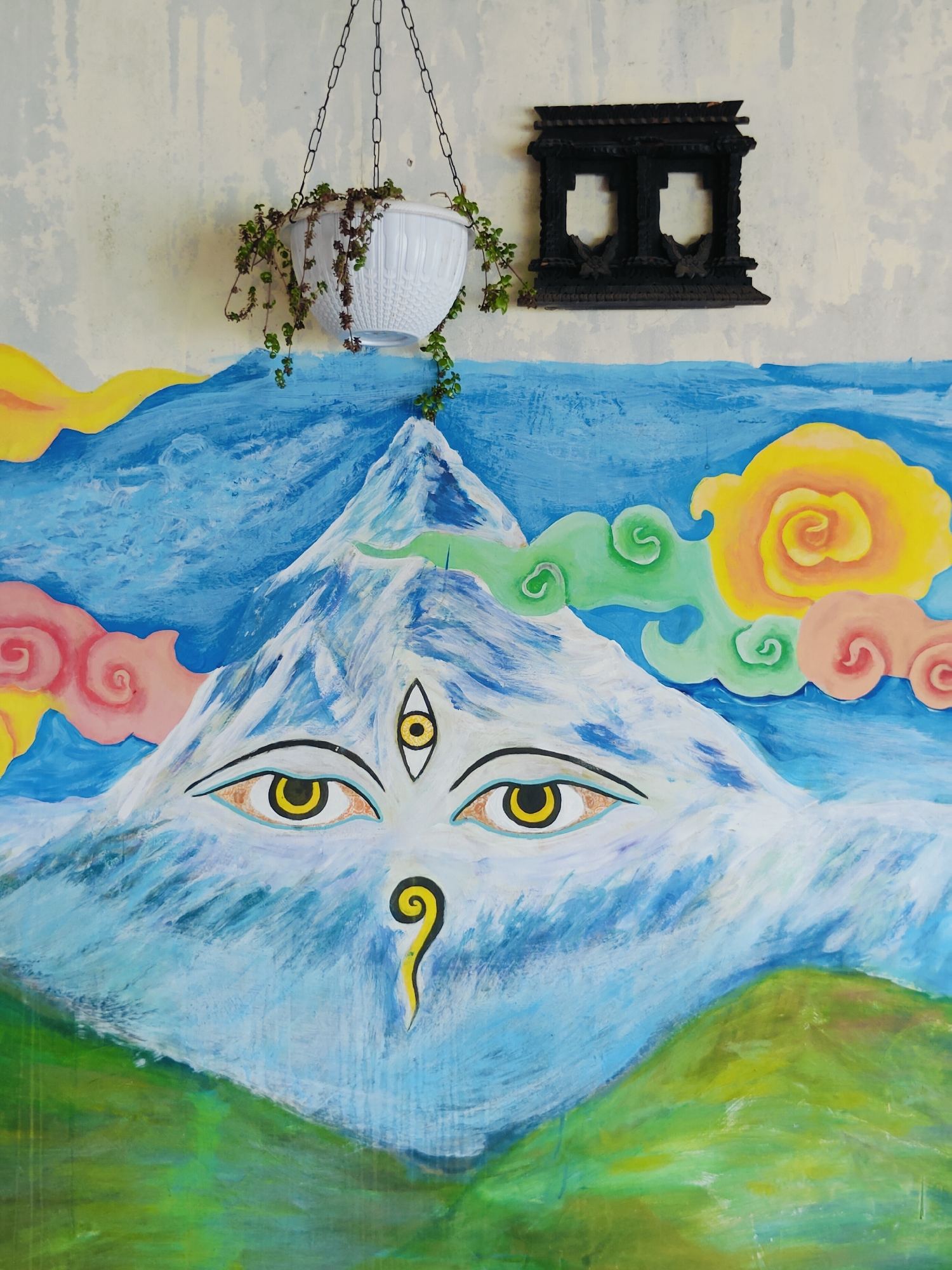 The image depicts a vibrant, close-up mural painted on a textured, light-colored wall. The mural's central subject is a snow-capped mountain with prominent, watchful eyes and a third eye on its forehead. The mountain is rendered in shades of white, blue, and hints of green, with streaks suggesting movement and texture. To the left and right of the mountain, stylized clouds in pink, yellow, and green swirl around it. The foreground of the mural features rolling green hills.
Above the mural, suspended by a black chain, is a white hanging planter containing a small, leafy green plant. To the upper right of the planter, a small, dark, intricately carved wooden object, resembling a decorative window frame, is mounted on the wall.
The wall itself has a worn, somewhat rough texture with subtle variations in color, appearing to be an exterior surface. The overall impression is one of artistic expression, possibly adorning a building or public space in Pokhara, Nepal, a city known for its stunning mountain views, particularly of the Annapurna range, which this mural might be referencing. The painting's style is somewhat naive and symbolic, evoking a sense of spirituality or cultural significance often associated with the region. There are no people or animals present in the scene.