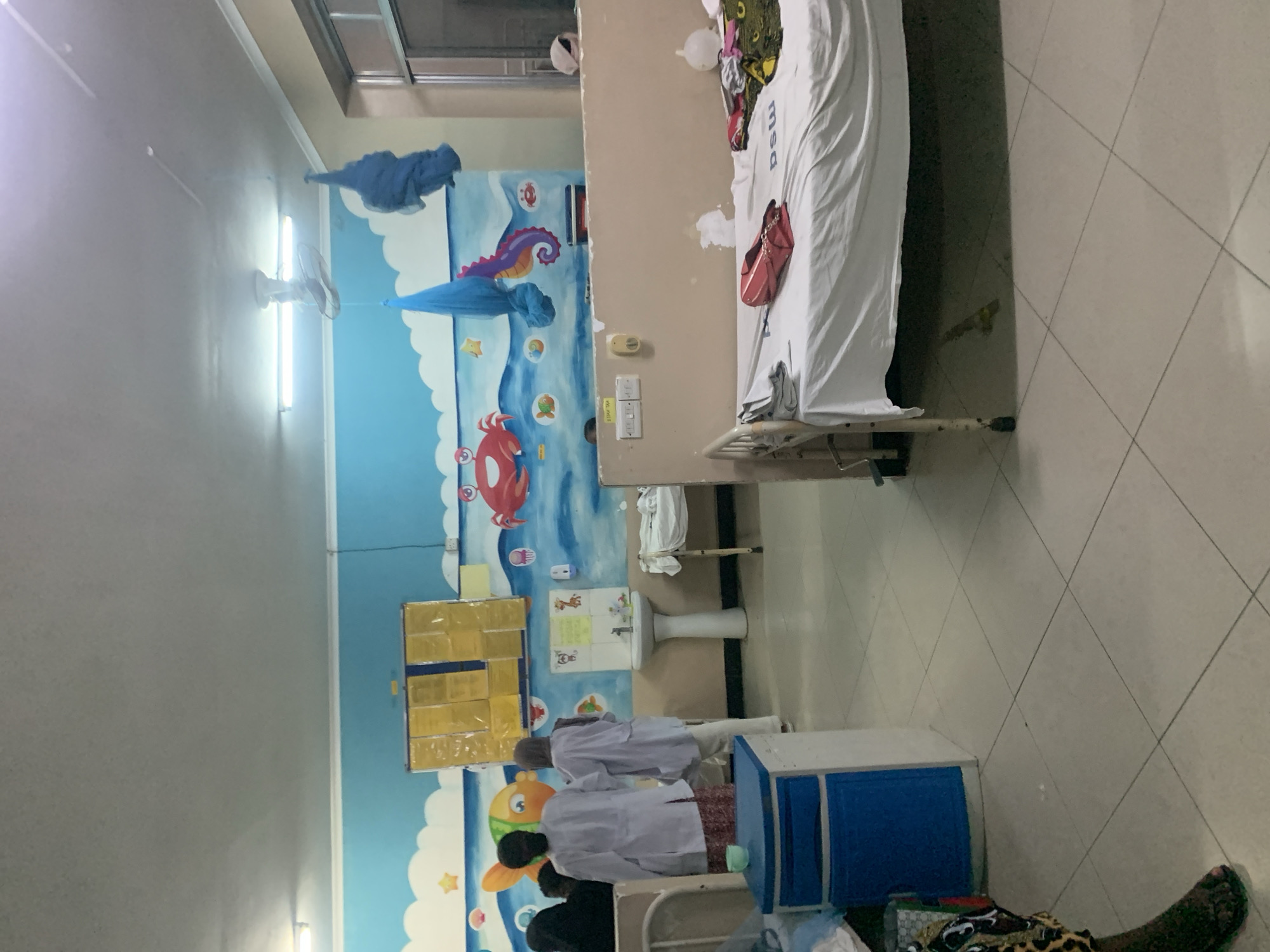 The image depicts a hospital room, possibly a children's ward, in Dar es-Salaam, Tanzania. The room features a bed with white linens, a blue bedside cabinet, and a sink. The walls are decorated with colorful murals of sea creatures. Two women wearing white coats are standing near the blue cabinet. One woman in a black shirt is beside them. The room has tiled floors, and a ceiling fan is visible.