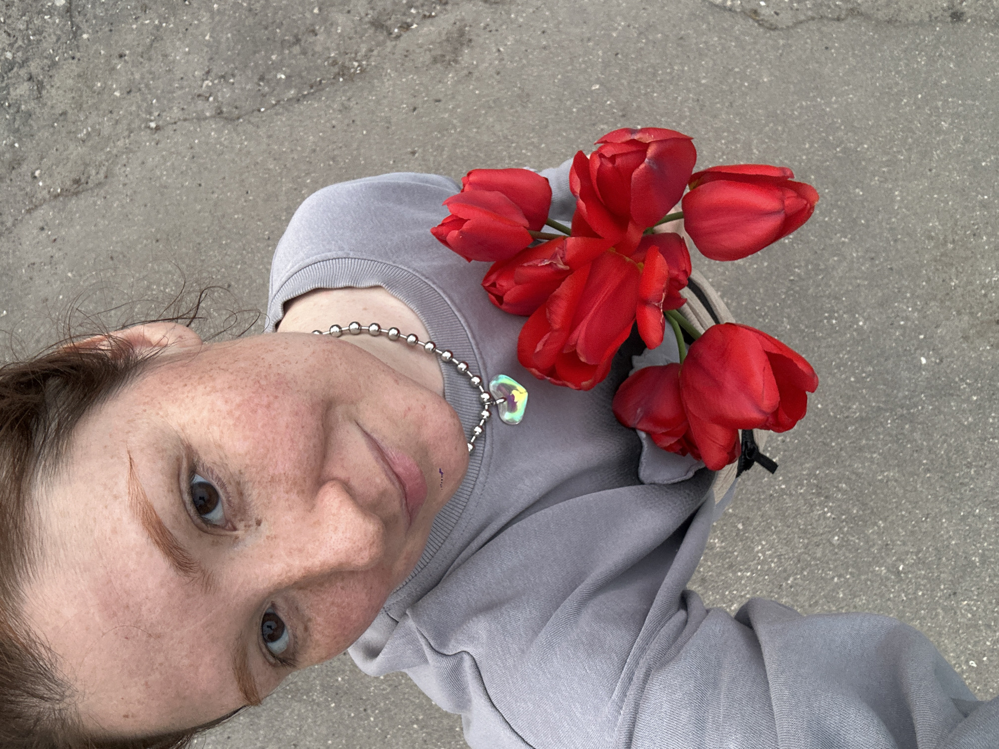 A person with freckles is holding a bouquet of red tulips. The person is wearing a gray sweatshirt and a silver necklace with a heart-shaped pendant. The background appears to be a gray paved surface. The image does not provide enough detail to identify if the location is in the city of Nizhny Novgorod, Russia.