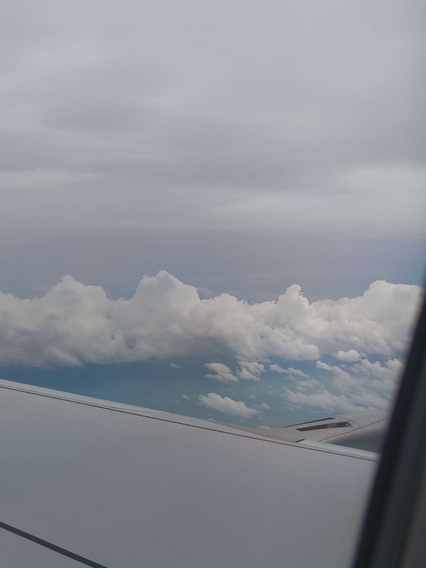 The image is a photograph taken from an airplane window, looking out at a cloudy sky. The lower portion of the image shows the wing of the airplane, which is light gray and smooth. Above the wing, there are layers of clouds. The clouds closest to the wing are white and fluffy, appearing to be cumulus clouds. Further up, the clouds become more diffused and gray, suggesting a layer of stratus clouds or an overcast sky. The overall lighting is muted, indicating it might be daytime with heavy cloud cover. There are no people or other identifiable objects visible in the scene other than the airplane wing and the sky. The image does not depict any specific location in the city of Giồng Kè, Vietnam.