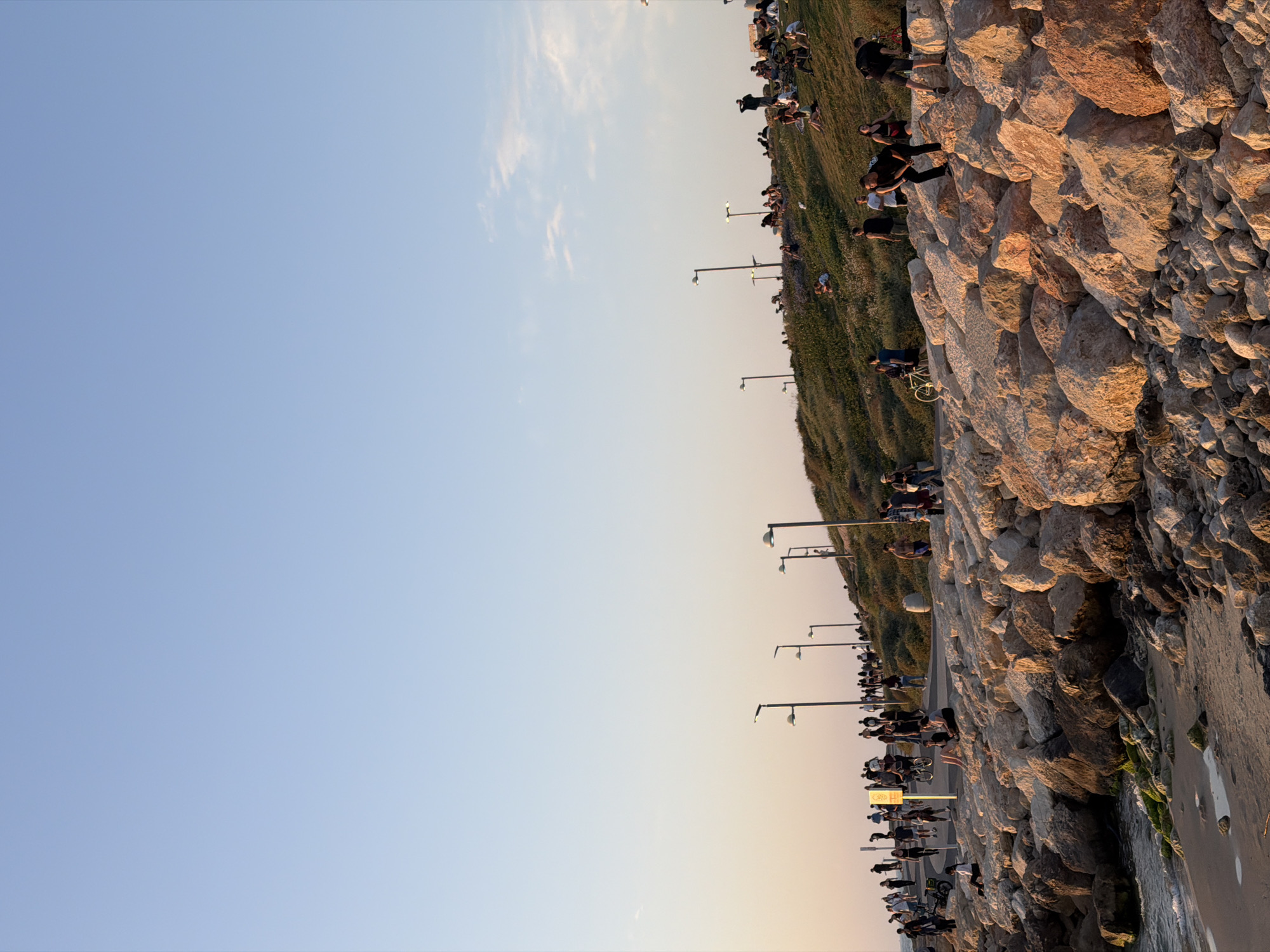 The image depicts a waterfront scene in Tel Aviv, Israel at sunset. The right side of the frame is dominated by a large, man-made rock barrier separating the promenade from the beach. People are sitting and standing along the top of the barrier. There are lamp posts extending along the walkway parallel to the barrier. The left half of the image is a clear sky with a gradient from light blue at the top to orange near the horizon. The people are engaging in casual activities such as walking, sitting, and cycling along the promenade. The overall atmosphere is relaxed and summery.