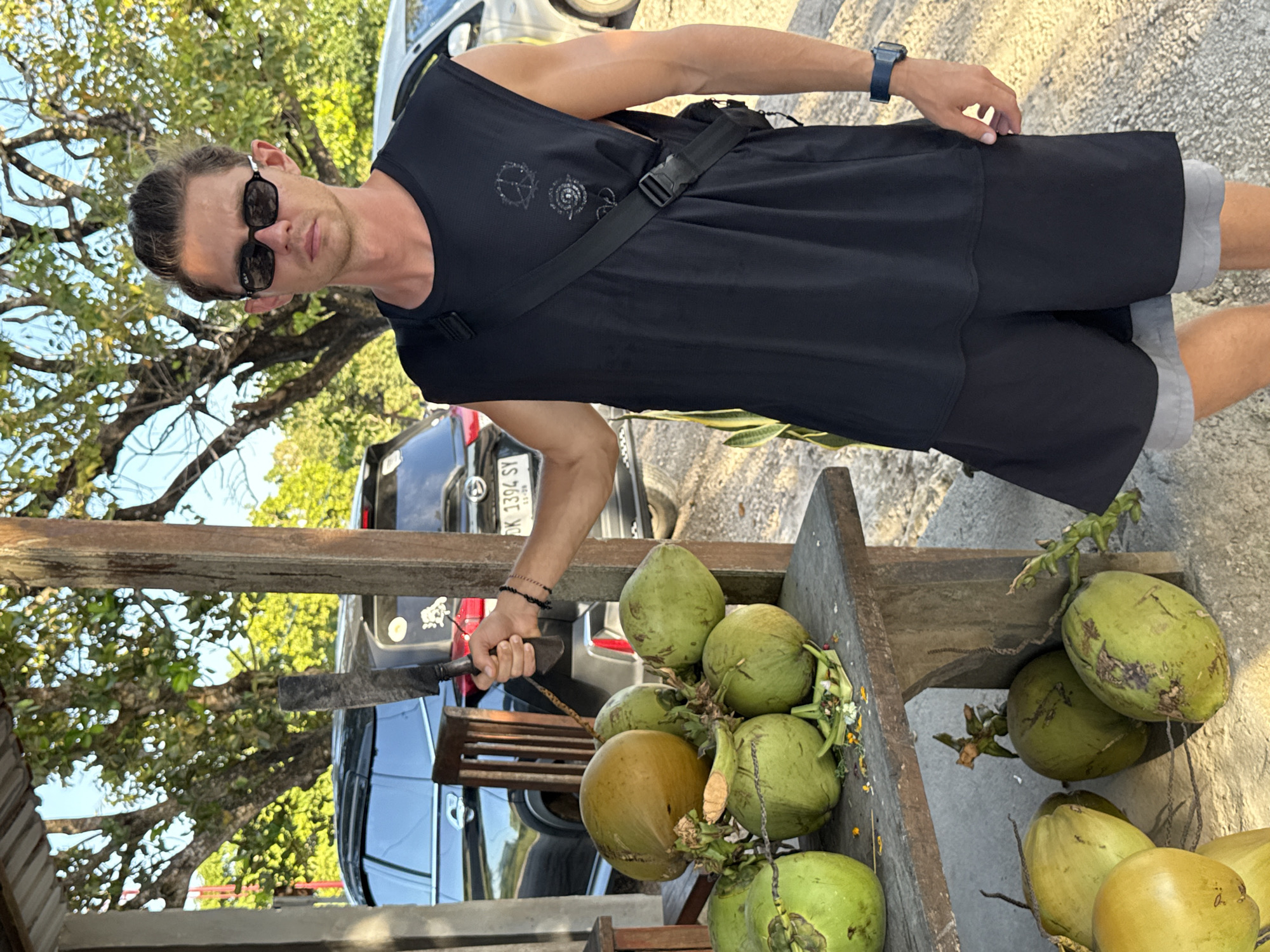 A man in sunglasses, a black sleeveless shirt, and shorts stands outdoors near a wooden structure laden with green and yellow coconuts. He holds a large knife. In the background, cars are parked, and lush green trees fill the sky. The location is likely Nusa Penida, Indonesia. The man appears to be preparing coconuts.