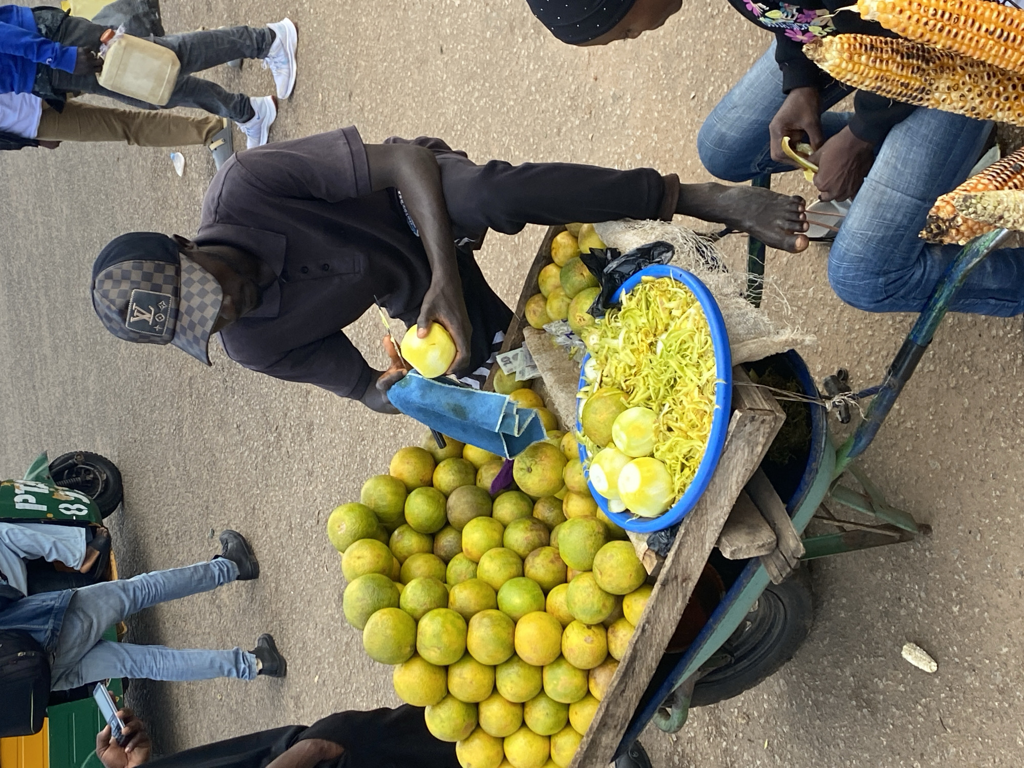 In Bukuru, Nigeria, a street vendor, a dark-skinned male wearing a black polo shirt and a checkered cap, sits barefoot beside a cart filled with oranges. He is in the process of peeling a yellow fruit, likely another orange, using a small knife. In front of him, a blue plastic bowl holds shredded yellow produce. A wheelbarrow, its wooden body filled with oranges and a blue plastic bowl, is positioned in the foreground. Two other individuals are partially visible; one, a person in blue jeans, sits and also appears to be peeling something, possibly corn on the cob, which is visible in the lower right corner. Another person, seen in the background, wears blue jeans and a black jacket and carries a white jerrycan. The environment is an outdoor, paved area, suggesting a street market setting.