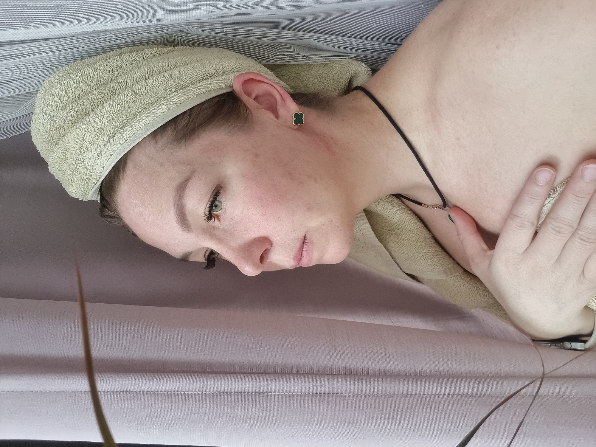 A medium shot depicts a person lying down, wearing a beige towel headband and a beige towel draped over their shoulder. The person has light skin, green eyes, and dark eyelashes. They are wearing a black choker with a pendant and clover-shaped earrings. Their left hand is resting on their chest. The background consists of a lavender-colored fabric and a striped white and gray fabric. A thin, green plant stalk is visible in the bottom left and right corners of the frame. The photograph appears to be taken indoors. No specific details suggest the location is Chaikovsky, Russia.
