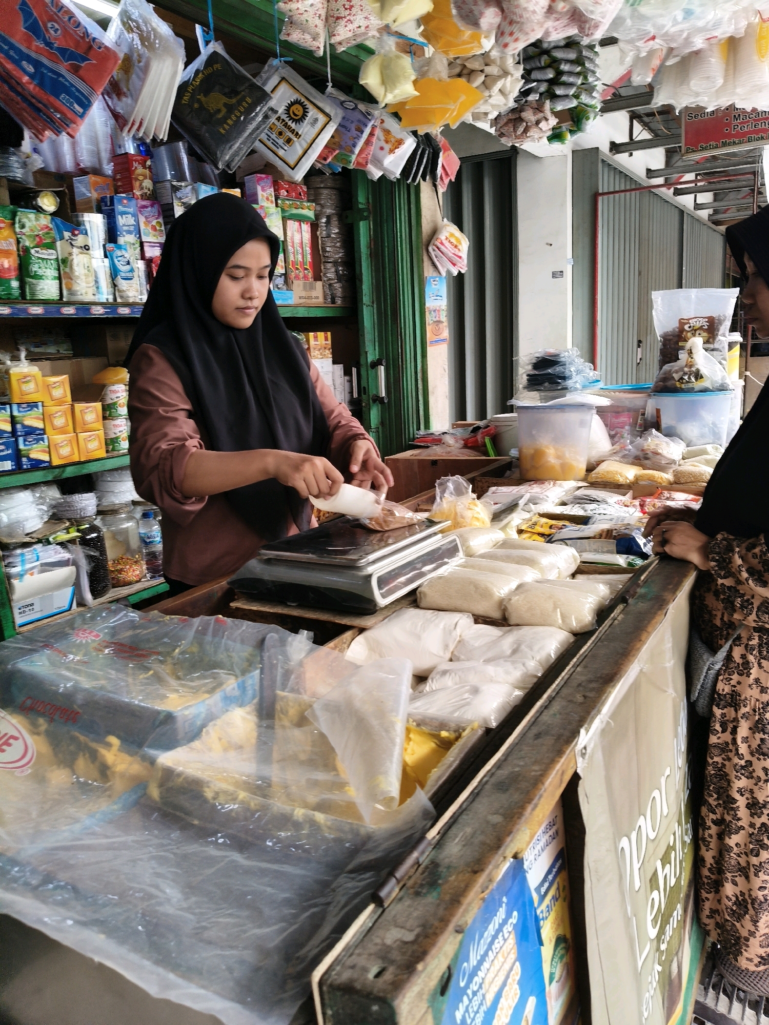 A young woman wearing a black hijab and a brown shirt is standing behind a market stall in Setiamekar, Indonesia. She is using a scale to weigh goods, holding a small white scoop in her right hand. The stall is filled with various products, including packaged food items, plastic bags containing rice, and other dry goods. The background shows more shelves stocked with products and a corrugated metal wall. To the right, another person, partially visible, is wearing a floral patterned dress. The stall appears to be an outdoor or semi-outdoor market environment.