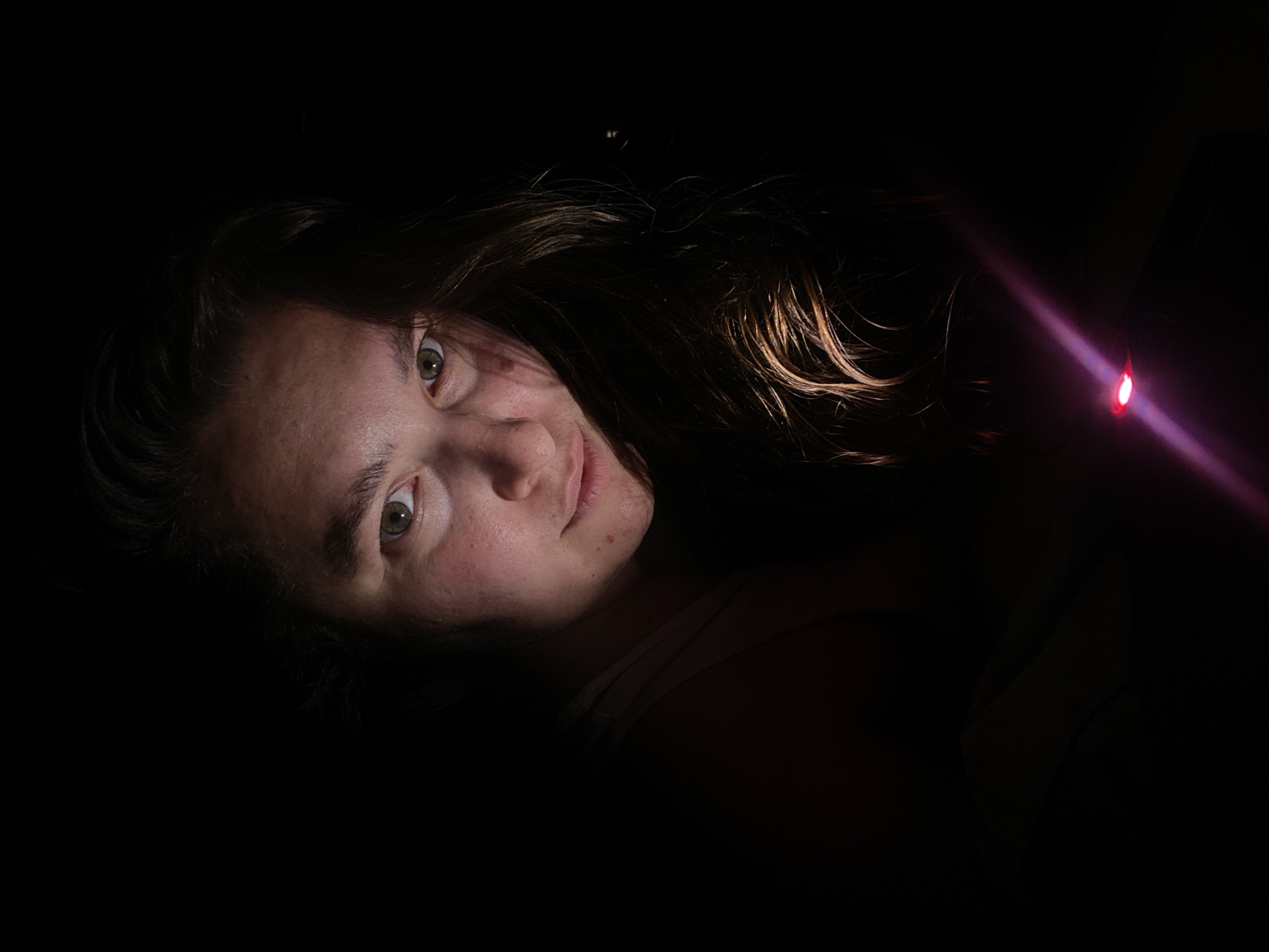 A close-up, low-key, portrait photograph of a young woman is captured against a dark background. She is lying down and looking directly at the camera. Her hair, a medium brown color, cascades around her face and shoulders. The lighting is dramatic, originating from below and to the right, illuminating her face from the side and highlighting strands of her hair with a warm, golden glow. A pinkish-purple light beam, likely from a phone or small flashlight, cuts diagonally across the lower right portion of the frame. Her skin is visible with subtle blemishes and natural texture. The overall mood is intimate and slightly mysterious due to the extreme darkness and focused lighting. The location in Perm, Russia cannot be determined from the image.