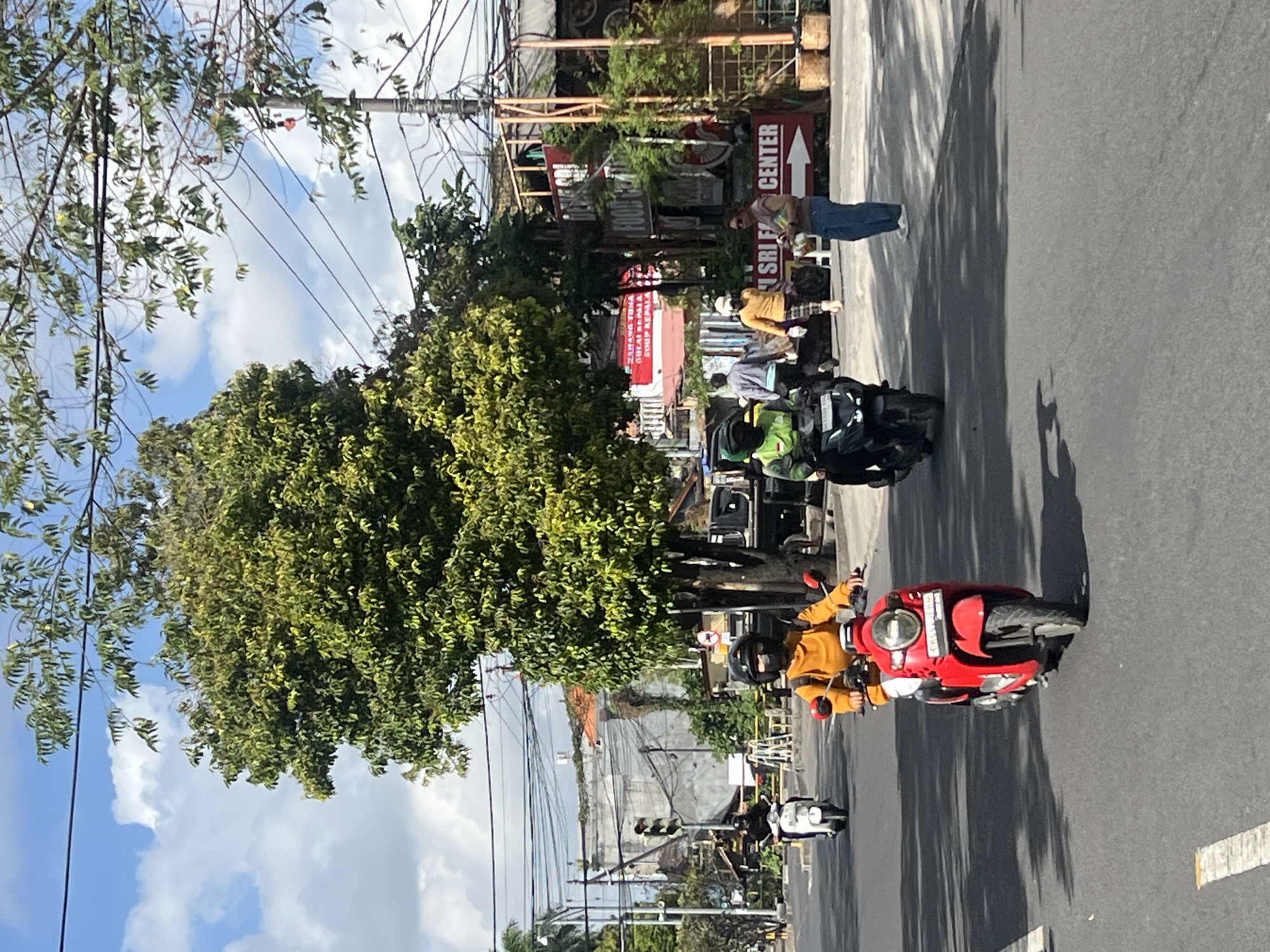 In Kuta, Indonesia, on a sunlit day with a partly cloudy sky, a street scene unfolds with multiple individuals riding motorcycles. In the foreground, a person wearing an orange shirt and a black helmet rides a red scooter, their face obscured by a black mask. Behind them, a person in a green helmet and jacket rides a black motorcycle. Further back, another person on a white scooter is visible. To the right, a woman in blue jeans and a brown top stands holding what appears to be a phone. A sign with red lettering reads "SRI FOOD CENTER". Electrical wires crisscross overhead, and lush green trees with dense foliage line the street. Buildings with red-tiled roofs are visible in the background. The road is paved, with white lane markings. Shadows are cast across the asphalt from the trees and overhead wires.