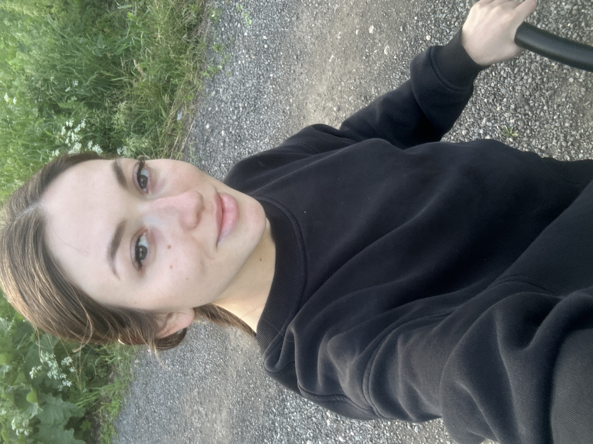 A young woman with brown hair and fair skin is taking a selfie outdoors. She is wearing a black crewneck sweatshirt. The background consists of a gravel path and lush green foliage. The overall lighting suggests it is daytime, likely late afternoon or early evening, with soft natural light. The location is Estachi, Russia.