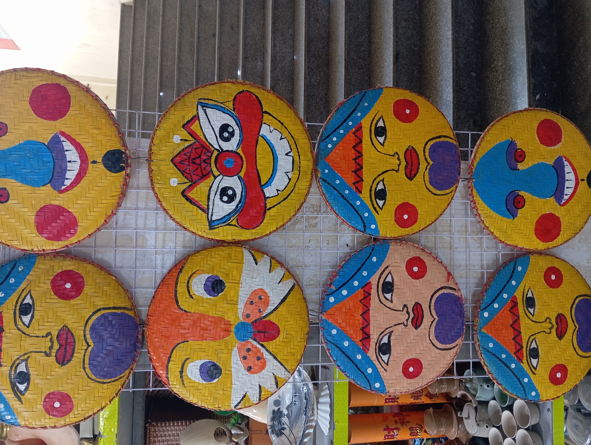 The image shows a display of woven bamboo masks, likely decorative or for sale, arranged in columns against a white grid. The masks are round and feature stylized faces painted in bright colors. They are all yellow with various features like large eyes, noses, mouths, and rosy cheeks. Some masks depict human-like faces, while others have more animalistic or monstrous features, like the one with a lion's mane and prominent fangs. The masks are hung on a metal grid, suggesting they are part of a market stall or shop display.
In the background, a concrete staircase is visible, indicating an indoor or semi-outdoor setting with multiple levels. To the left of the masks, there are glimpses of other items, including what appear to be ceramic cups and teapots, as well as cylindrical orange objects with red Chinese characters, potentially incense sticks or decorative tubes. The lighting suggests it is daytime.
While the specific location within Nha Trang is not explicitly identifiable from the image alone, the presence of the bamboo masks and potentially Vietnamese or Chinese characters on some items strongly suggests a cultural or tourist-oriented market in Vietnam. Many tourist markets in Nha Trang sell similar handicrafts.