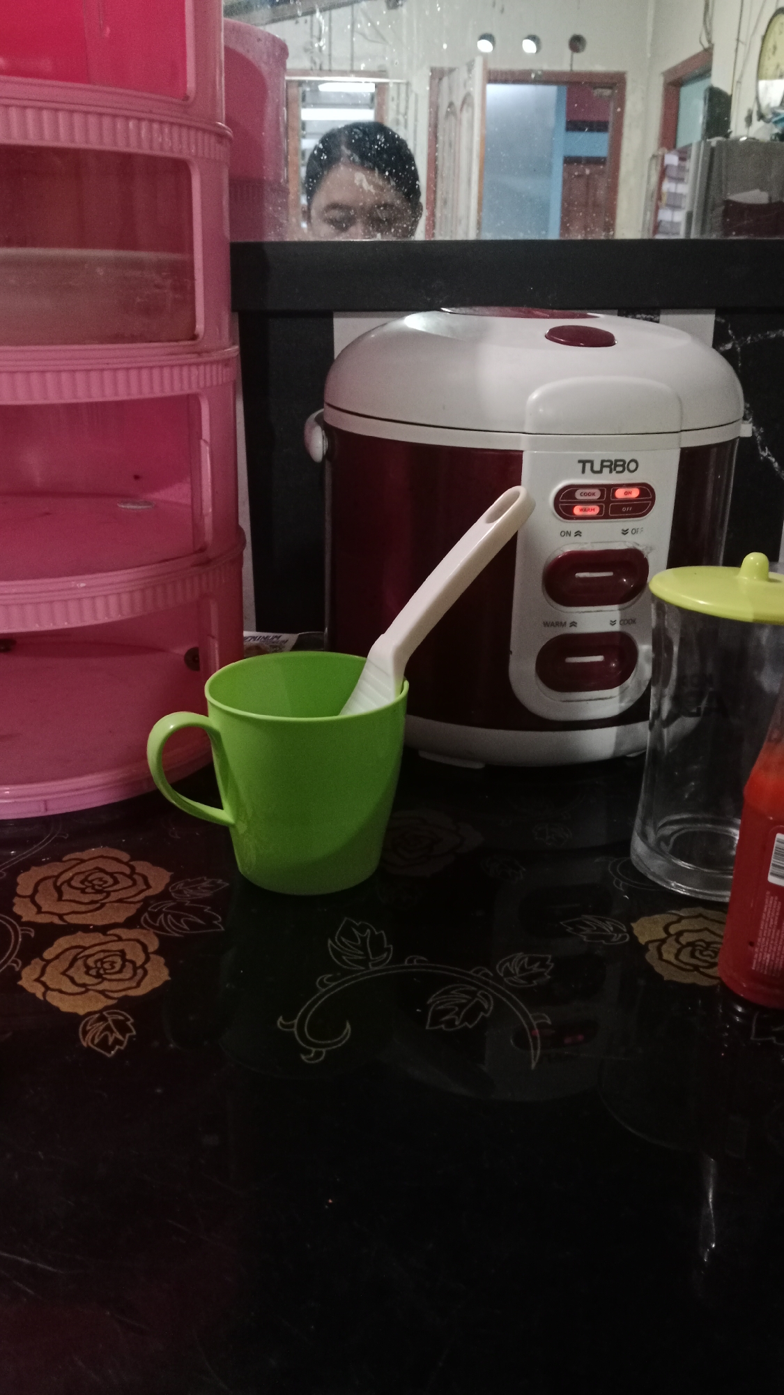 The image captures a domestic scene, likely a kitchen counter, in Blitar, Indonesia. A pink tiered storage unit is on the left, reflecting a person's face, partially obscured, in a mirror above the counter. In the center, a red and white Turbo brand rice cooker is prominent, with its control panel displaying "COOK" and "ON" illuminated. To the right of the rice cooker, a clear glass jar with a lime green lid sits next to a red bottle, possibly a sauce. A bright green mug containing a white utensil is placed in front of the rice cooker. The counter surface itself is black and decorated with floral patterns.