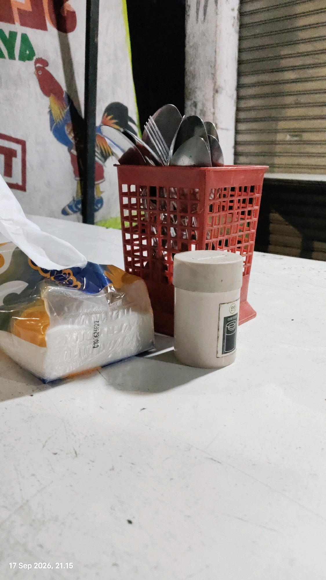 The image depicts a close-up view of a white table with various items, likely in an outdoor dining setting in Surabaya, Indonesia, given the timestamp. In the foreground, a package of tissues is prominently displayed, showing a partially opened plastic wrapper with a white tissue pulled out. To the right of the tissues, there is a small, cylindrical beige container with a light-colored lid, featuring a label with a circular logo and some text. Behind these items, a red plastic cutlery holder is filled with spoons and forks. The background shows a portion of a wall with a mural of a rooster and abstract shapes, typical of street art or signage in Indonesia. Further in the background, a gray roller shutter door is visible, suggesting a storefront or establishment. The overall lighting is dim, creating shadows and highlighting the textures of the table and objects. The timestamp in the bottom left corner indicates "17 Sep 2026, 21:15".