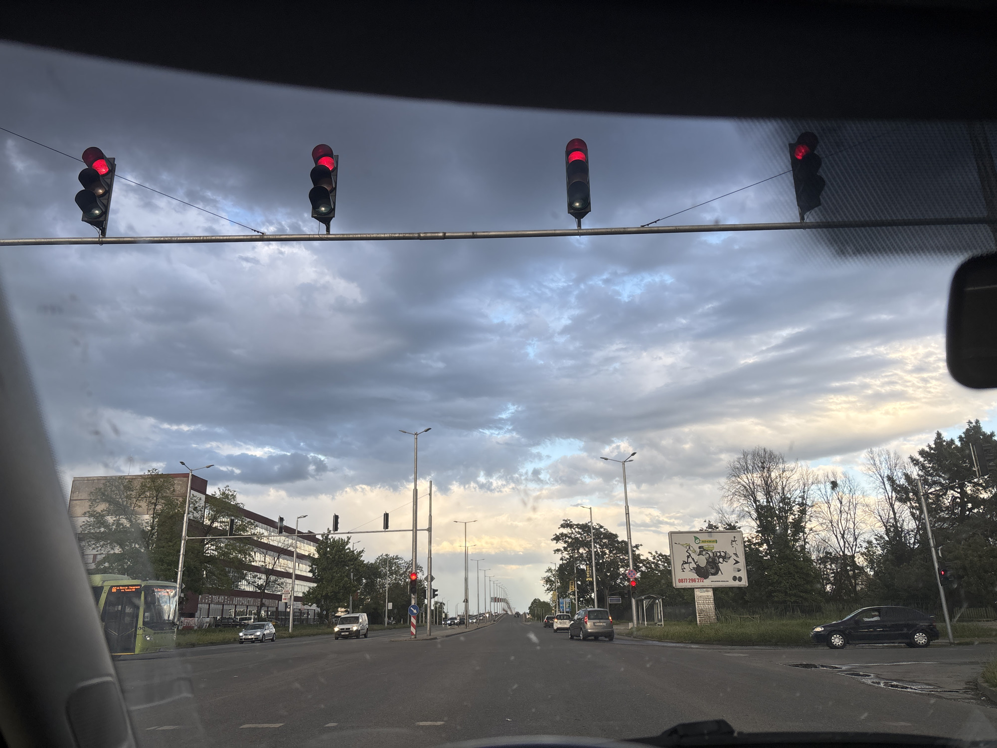 The image depicts a view from inside a vehicle, likely a car, looking out onto a city street. The sky is overcast with heavy, dark clouds. Traffic lights, all displaying red, are suspended above the road. The road is wide with multiple lanes and streetlights lining both sides. Several vehicles, including a bus on the left side of the road, are present. In the background, there is a building and trees. The scene appears to be in an urban area of Plovdiv, Bulgaria, based on the visual elements present. There are no people visible in the image.