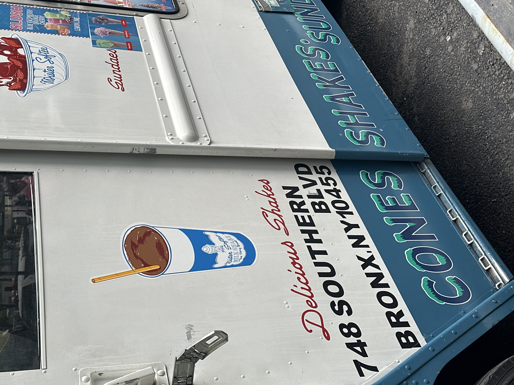 The image captures the side of a white and blue ice cream truck, parked on an asphalt surface under bright daylight. The truck is covered with vibrant graphics and text detailing its offerings.
On the upper white section, a graphic features a bowl of strawberries with syrup, labeled "Mister Softee" and "Sundaes" in red cursive. Adjacent to this, a "SLUSH" menu advertises "BLUE RASPBERRY," "ORANGE," "CHERRY," and "LEMON LIME" flavors, along with images of colorful slush drinks and ice cream cones (Strawberry, Lime, Pistachio).
Further down the white panel, a large illustration depicts a chocolate milkshake in a cup with a straw. This cup bears a "Mister Softee MILK SHAKE" logo, and the phrase "The Very Best Mister Softee" is also visible in the branding. Beside the shake graphic, the text "Delicious Shakes" is written in elegant red cursive. Below this, the truck's address is clearly displayed in black: "748 SOUTHERN BLVD BRONX.NY 10455".
The lower part of the truck features two distinct blue panels. The upper blue panel prominently displays "SHAKES SUNDAES" in large, outlined green and blue letters. Directly below, the second blue panel reads "CONES" in the same distinctive lettering style.
The overall scene presents a mobile vendor of frozen desserts, showcasing its brand, menu, and location clearly. No people are visible in the frame.