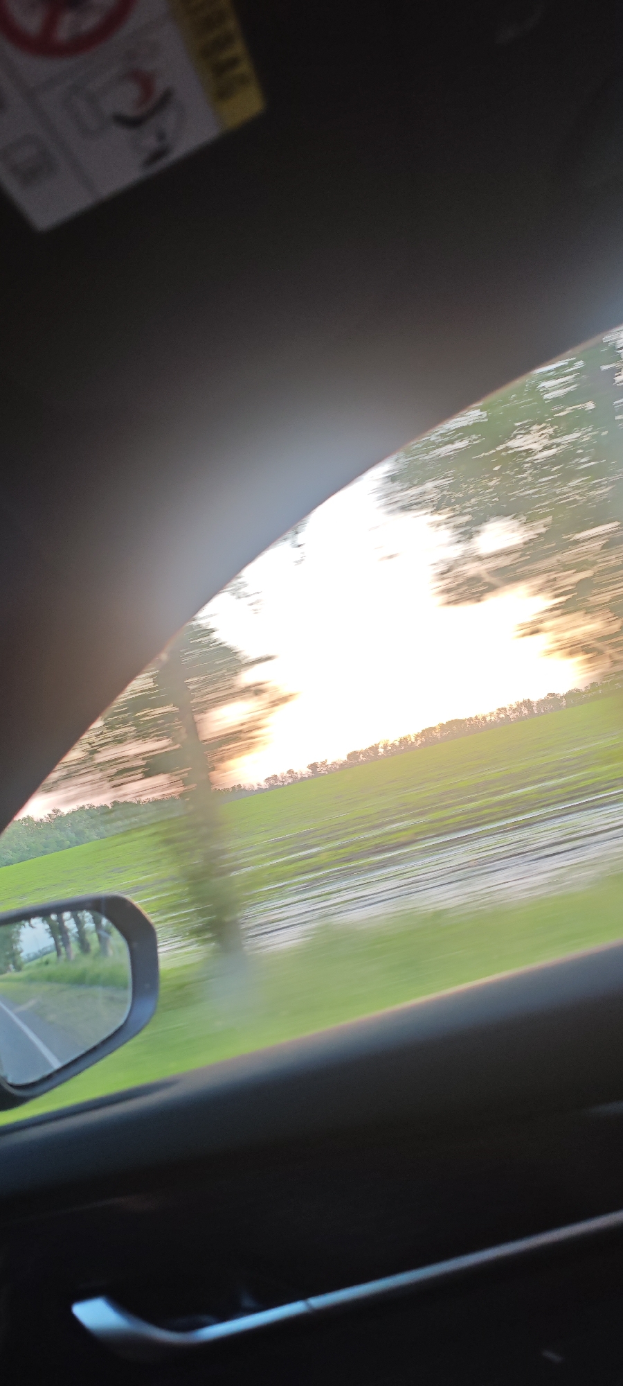 The image is taken from inside a moving vehicle, likely a car. The view is through the passenger window. Outside the window is a rural landscape with a field or pasture that appears to be flooded or very wet. There is a tree line in the distance. The sky is bright, possibly indicating sunrise or sunset. The side mirror of the car is visible, showing a road and some trees. The interior of the car, including the door panel and handle, is visible. An airbag warning label is partially visible at the top of the frame. The scene appears to be in motion due to the motion blur. There is no indication of the city of Beysug, Russia.