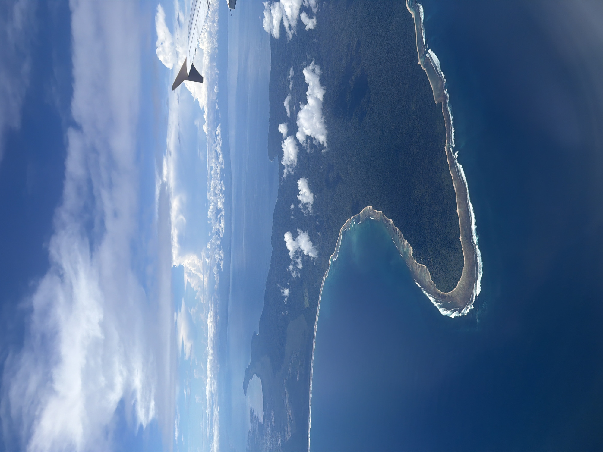 An aerial photograph captures a breathtaking view of a tropical coastline and ocean from an airplane, likely near Denpasar, Indonesia. The perspective is from above, looking down and forward.
In the upper left corner, a portion of an airplane wing is visible, featuring its distinctive wingtip. The sky, dominating the upper section of the image, is a vibrant blue adorned with numerous fluffy white cumulus clouds. Towards the horizon on the left, the sky appears hazier.
Below the sky, the vast expanse of the deep blue ocean stretches out, meeting a richly forested landmass in the foreground. This landmass, likely an island or peninsula, is covered in dense, dark green tropical foliage. Its coastline is well-defined by a narrow strip of white, representing sandy beaches or coral reefs, where bright white waves are seen breaking. The coastline exhibits a distinctive curved shape, particularly prominent on the right side, resembling a hook or crescent.
Further inland on the main landmass, a lighter blue body of water, possibly a lake or lagoon, is discernible amidst the greenery. Scattered white clouds cast shadows or drift gently over the landmass. The overall impression is one of a clear, sunny day, providing excellent visibility of the stunning natural landscape below. No people or visible text are present in the image.