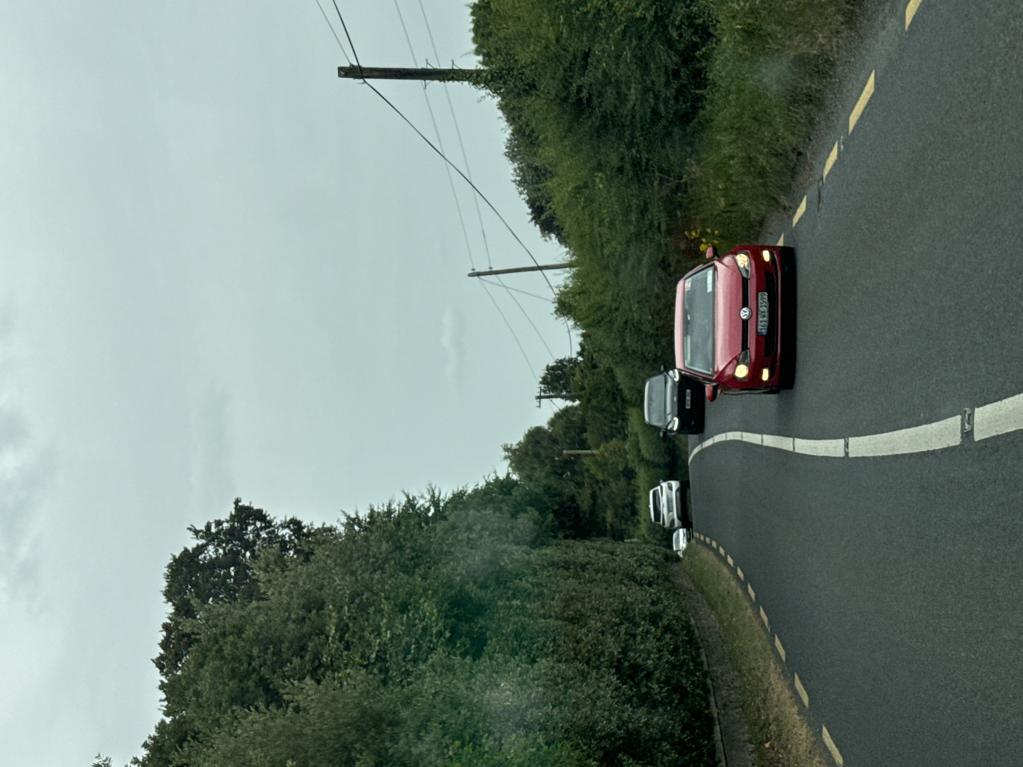 The image shows a view from a vehicle driving on Eyrefield Road in Ireland. The road is asphalt and has a white dashed line and a solid white line separating lanes. The road is bordered by thick green hedges and trees on the left and right. In the distance, there are several cars on the road. The sky is overcast and gray. The red Volkswagen Golf in the foreground has the license plate 161-WX-1599. A dark-colored SUV is behind it, and further ahead, a white SUV and another white car are visible. Utility poles with wires are present on the right side of the road.