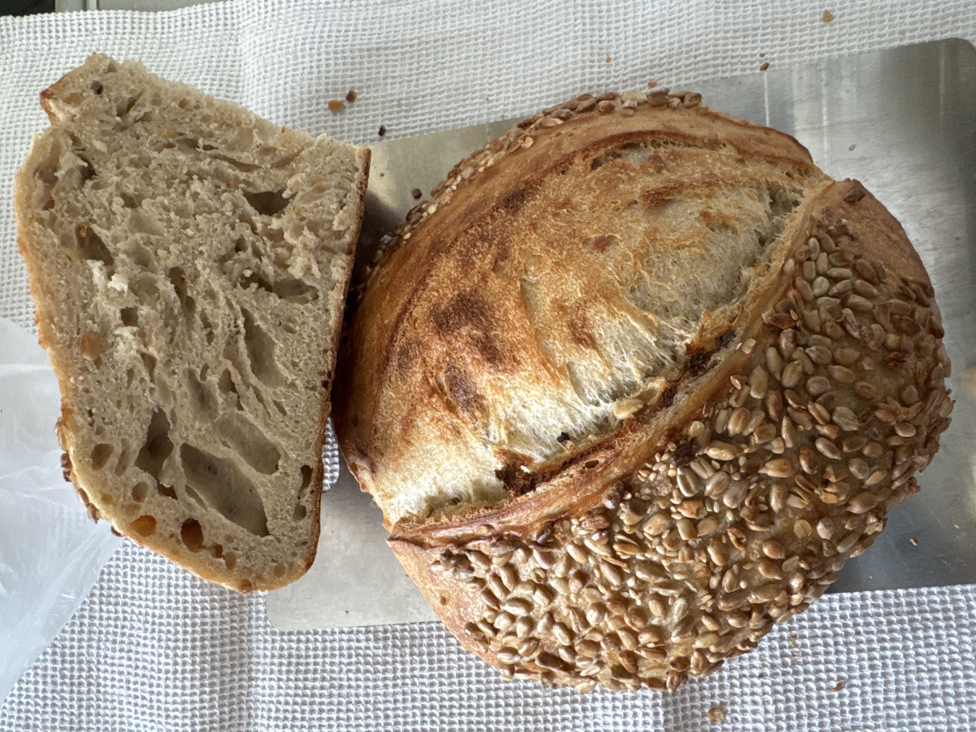 A slice of sourdough bread, with an open crumb structure, rests next to a round loaf of sourdough bread, coated with sunflower seeds, on a metal tray. The scene takes place on a white, textured tablecloth. The location is Высокий, Russia.