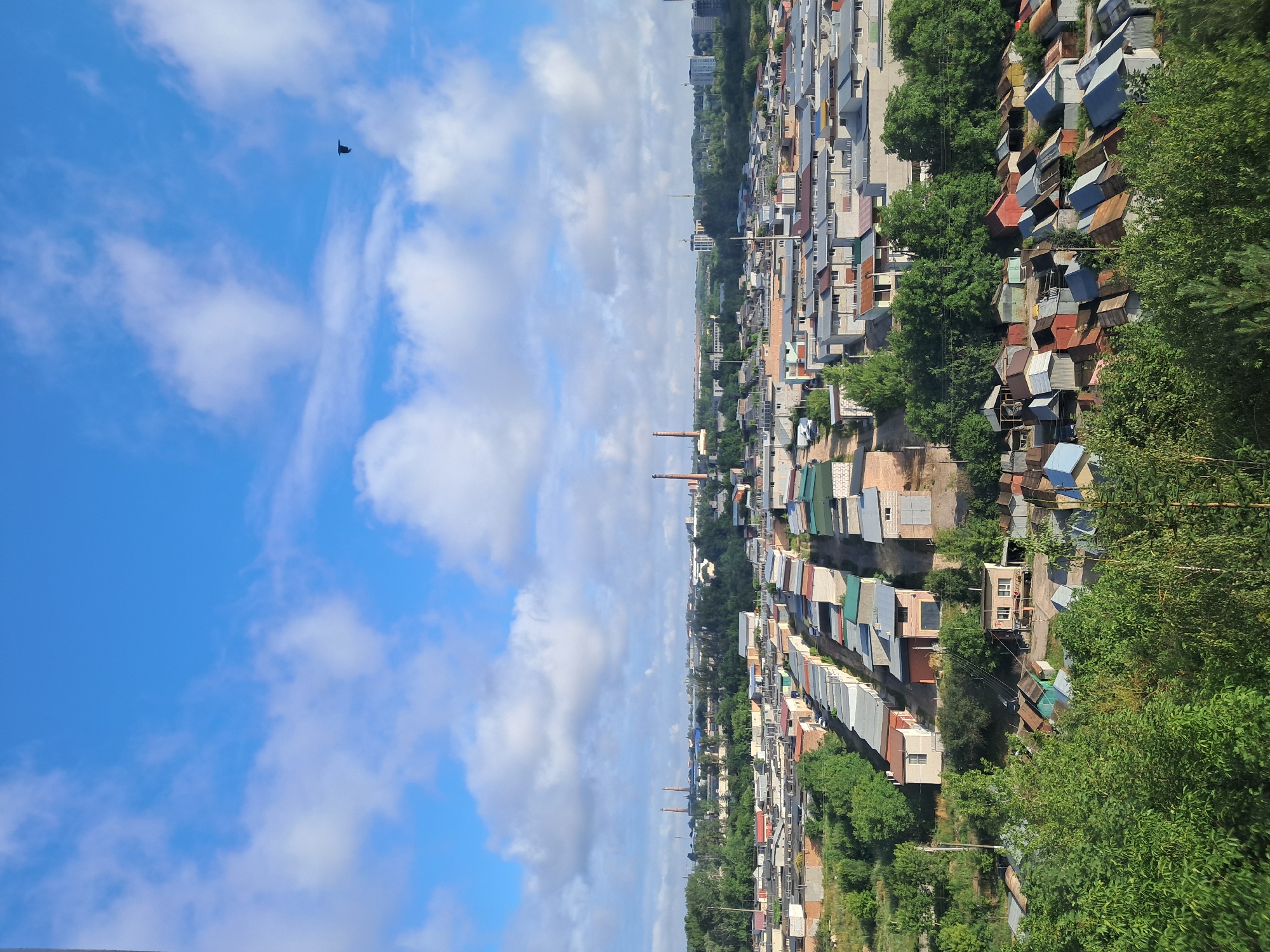 An aerial view captures a dense urban landscape filled with numerous garages and small buildings, likely a residential or industrial area. The scene is dominated by a vast expanse of tightly packed garages, many with metal roofs in various colors, including grey, red, and blue. Some of these structures appear weathered, with some roofs showing rust or damage. In the background, a more distant cityscape is visible, featuring taller buildings and industrial chimneys that punctuate the horizon. The environment is characterized by a clear blue sky with scattered white clouds, suggesting fair weather. Lush green trees and foliage are interspersed throughout the scene, particularly in the foreground and between some of the garage rows, indicating a blend of built environment and natural elements. A single bird is visible in flight against the sky. The overall impression is one of a working-class urban or suburban neighborhood with extensive private storage facilities.