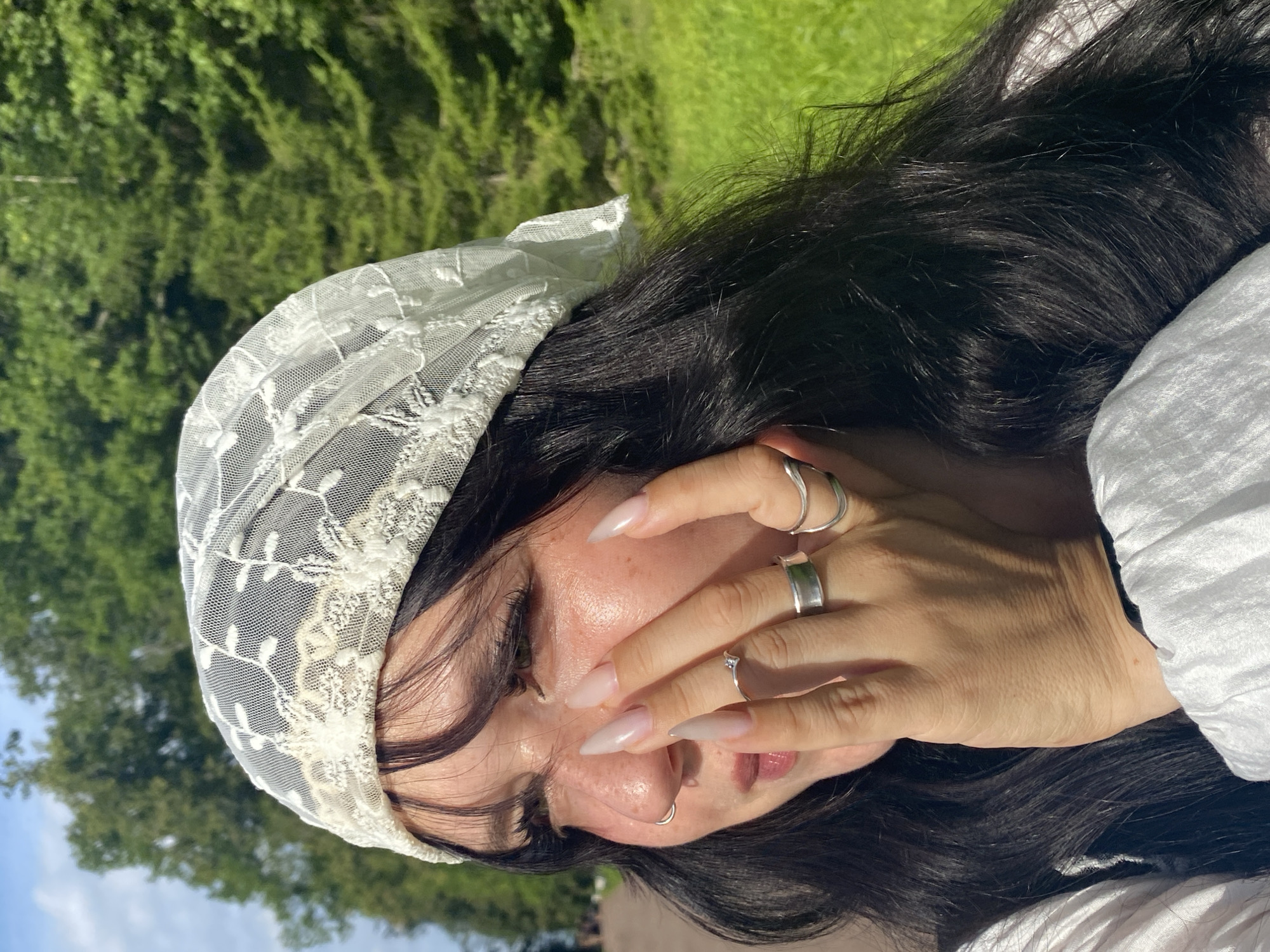 In this close-up, outdoor portrait, a young person with long, dark, wavy hair and bangs is positioned against a backdrop of lush green trees and a bright blue sky. They are wearing a white lace headscarf with an intricate floral pattern and a white collared shirt. The individual is looking to their left, with their right hand near their face, fingers adorned with several silver rings and long, light-colored artificial nails. A silver nose ring is visible on their right nostril. The person's face, partially obscured by their hand, shows visible freckles and is illuminated by natural sunlight, casting gentle shadows and highlighting their features. The overall setting suggests a warm, sunny day in a natural, possibly park-like environment, consistent with areas within the city of Lomonosov, Russia, known for its numerous parks and green spaces.