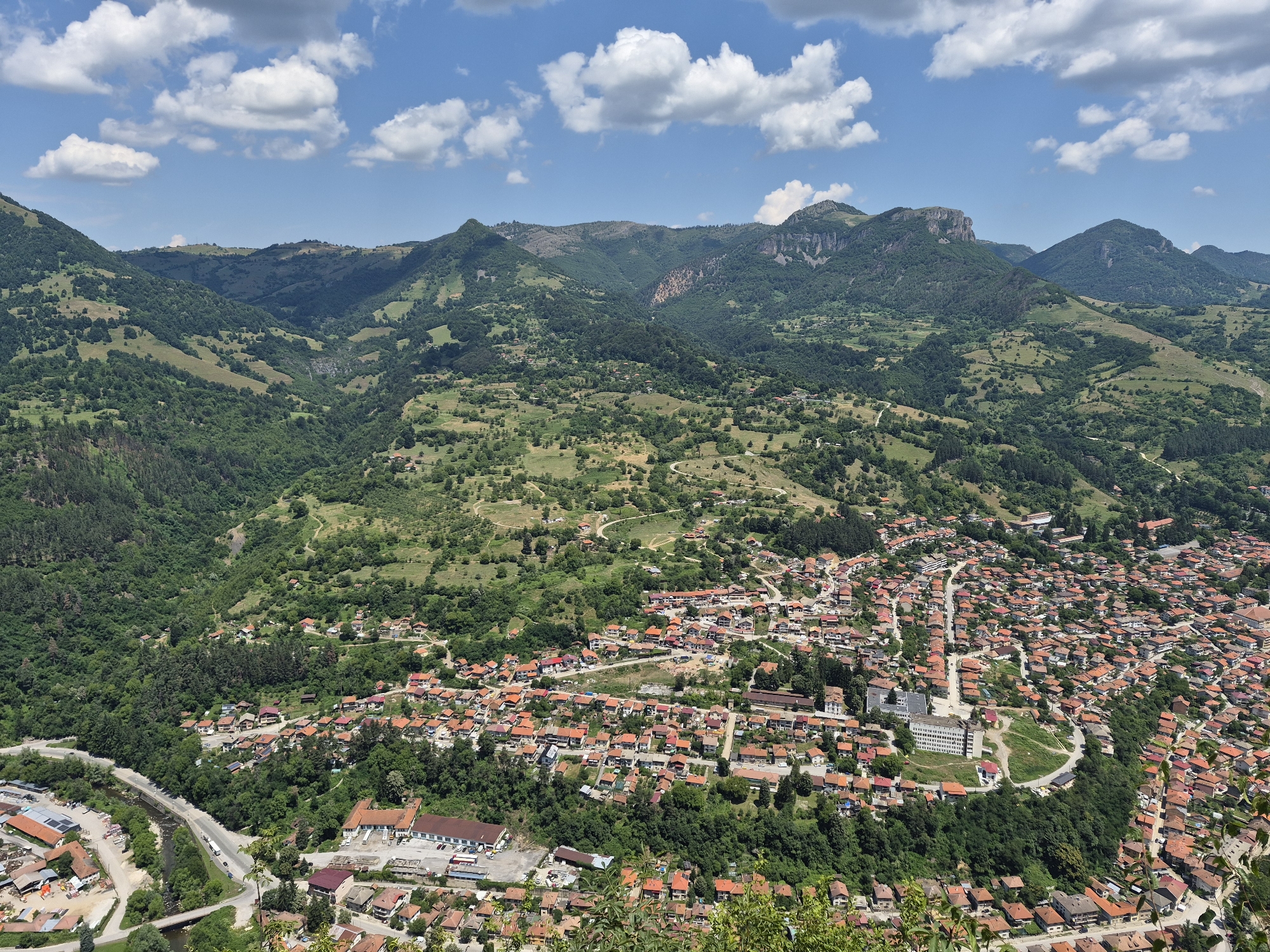 This is a high-angle aerial view of the city of Teteven, Bulgaria, on a clear day with scattered white clouds in a blue sky. The city is nestled within a lush green valley, surrounded by rolling hills and larger, verdant mountains in the background. The architecture of Teteven is characterized by numerous houses with red-tiled roofs, tightly packed together, with some larger buildings and structures interspersed. Roads wind through the urban area and connect to a larger road that runs alongside a river or stream on the left side of the image. The natural environment is a dominant feature, with dense forests covering the hillsides and mountains, and smaller patches of green spaces and cultivated land visible within and around the city. No people are discernible in this wide-angle shot. The overall scene depicts a scenic, relatively small city integrated into a picturesque mountainous landscape.