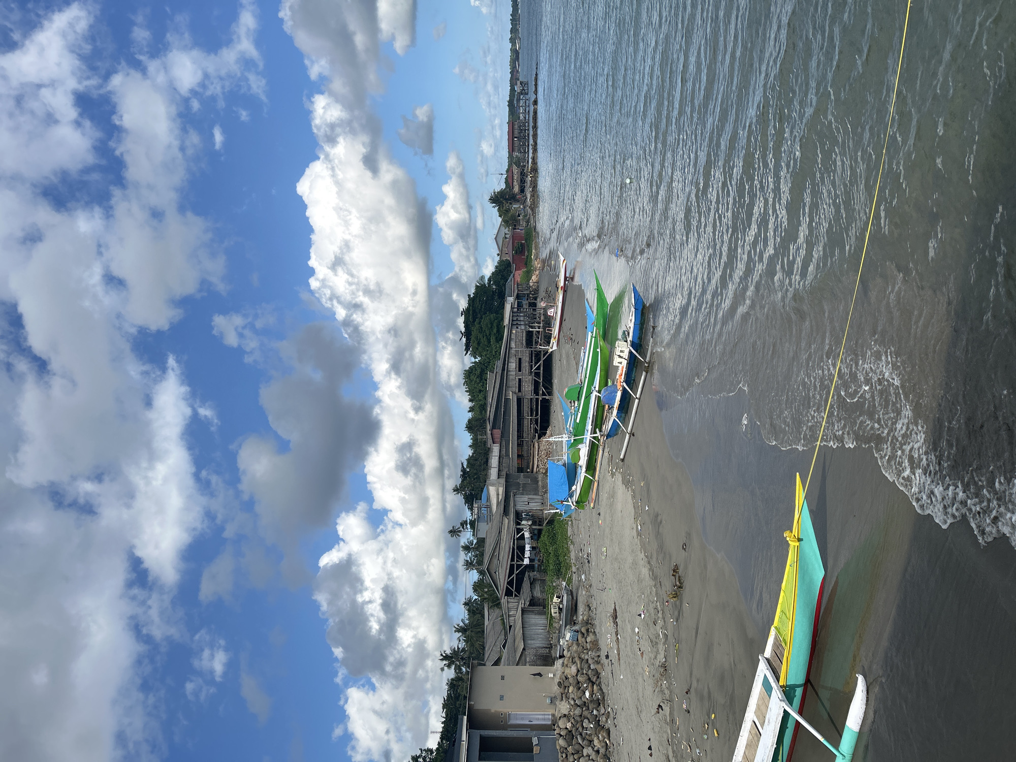 This image depicts a tropical beach scene in Ara, Indonesia. The foreground features several colorful outrigger boats pulled up onto the sandy shore. These boats are predominantly green and blue, with some accented with red and yellow. The water is a calm, dark blue, with gentle waves lapping at the beach. A yellow rope is visible extending from one of the boats into the water.
Behind the boats, a small coastal village lines the shore. The buildings appear to be constructed from wood and tin, with some showing signs of age and weathering. Palm trees are scattered throughout the village, adding to the tropical ambiance. In the background, the horizon is a mix of green vegetation and distant structures.
The sky is a vibrant blue, dotted with large, fluffy white clouds that cast shadows on the landscape. The lighting suggests it is daytime, likely mid-morning or afternoon. The overall mood is peaceful and serene, capturing the essence of a quiet seaside community. No visible text is present in the image.