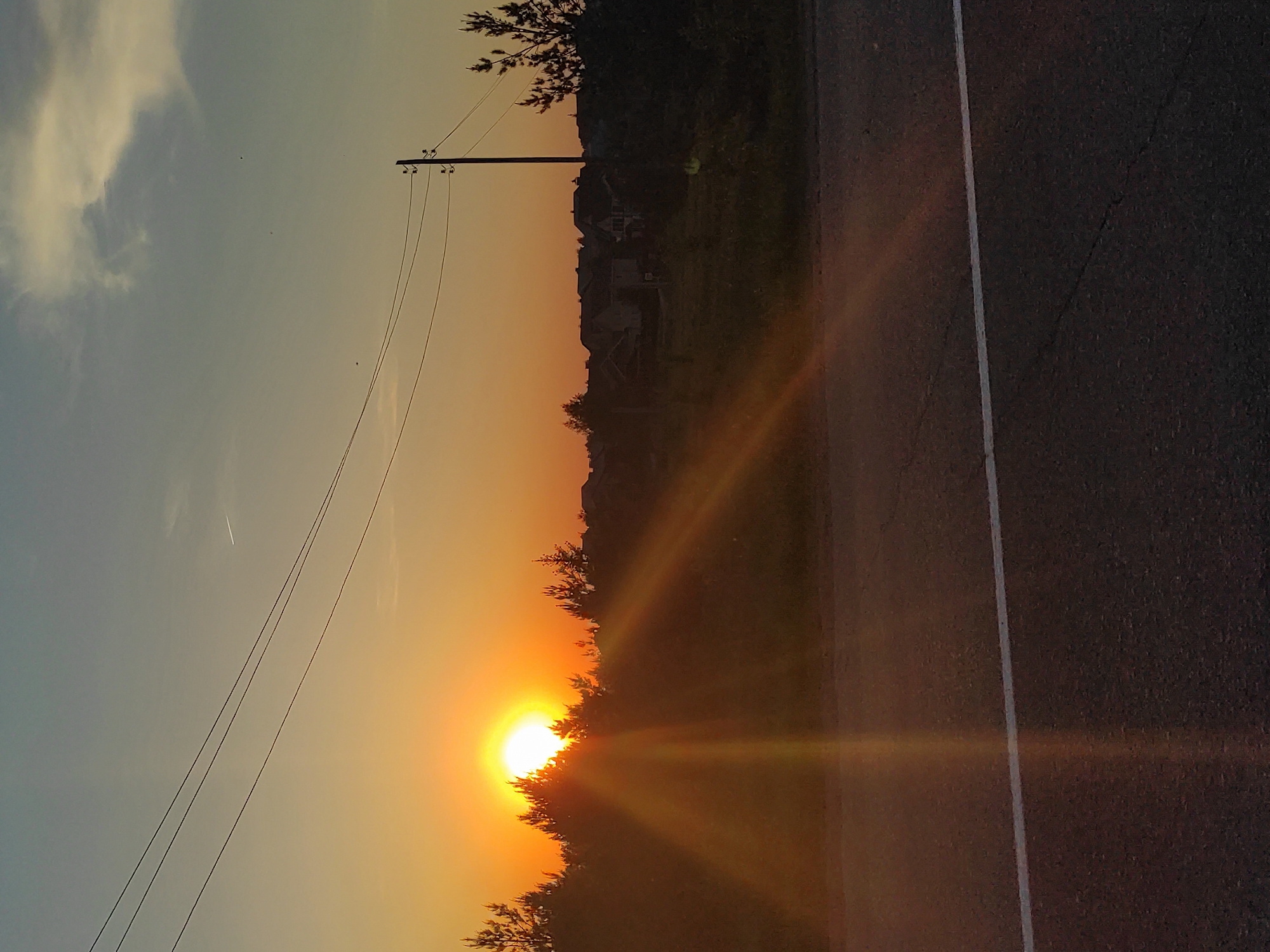 A photograph captures a sunset over a road in Прикамский, Russia. The sun is setting behind a line of trees, casting golden rays across the asphalt. An electrical pole and wires are visible on the left side of the frame. The sky is a gradient of blue to orange. A white line marks the edge of the road.