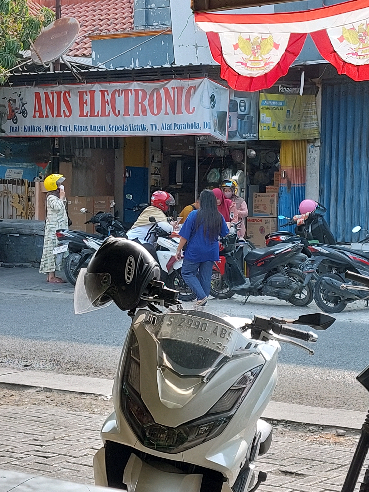 The image depicts an outdoor scene in front of an electronics store in Sumuragung, Indonesia. A banner above the store reads "ANIS ELECTRONIC" and lists items for sale including refrigerators, washing machines, fans, electric bicycles, televisions, and satellite dishes. Several motorcycles are parked in front of the store.
In the foreground, a white motorcycle with a black helmet on its handlebars is partially visible. The helmet has a clear visor and the brand name "INK" is visible on it. The license plate of the motorcycle reads "S 2990 AB5 03-24".
Behind the foreground motorcycle, several people are interacting. One woman wearing a yellow helmet and a long, patterned dress is standing on the left, facing away from the camera. Another woman in a pink headscarf and a light pink outfit is speaking to someone obscured by the motorcycle in the center. A person in a blue shirt and pants is standing near the motorcycles. Another individual in a red helmet and a light-colored shirt is sitting on a motorcycle to the right. A person with a pink helmet is sitting on a motorcycle further to the right.
The store entrance is visible with various items displayed, including what appear to be appliances like fans. Red and white Indonesian flags are hung above the store. The environment is a street with pavement in the foreground and a road beyond. The day appears to be sunny, casting shadows.