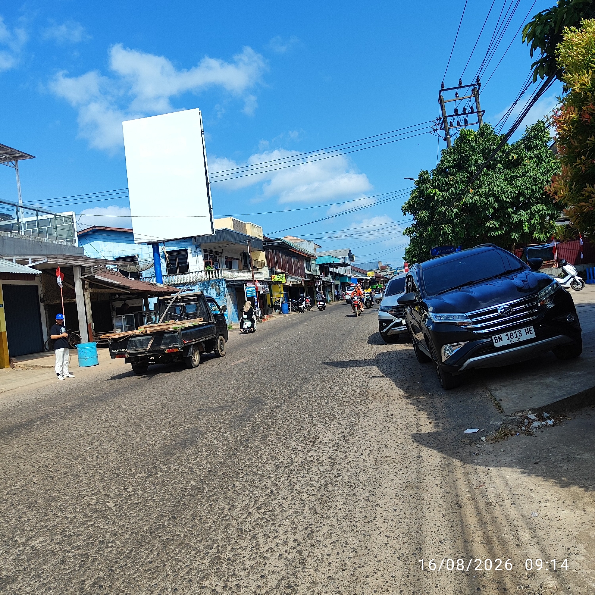 The image captures a street scene in Pembaharuan, Indonesia, on August 16, 2026, at 09:14. The foreground is dominated by a rough asphalt road with a noticeable texture. On the right side of the road, a black SUV, identified by its license plate "BN 1813 WE", is parked or stopped, casting a long shadow across the pavement. To the left of the SUV, another vehicle, possibly silver, is partially visible. Further down the street, a black pickup truck, labeled "SUZUKI CARRY", is loaded with lumber.
A person wearing a blue cap and white pants stands on the left side of the road, near a blue barrel and a small Indonesian flag on a pole. The background features a row of buildings with various architectural styles and colors, including blue and beige structures, some with tin roofs. Overhead, electrical wires are strung across utility poles. The sky is bright blue with scattered white clouds, indicating a clear day. Trees, some with green foliage and others with reddish-brown new growth, are visible along the right side of the street. Several motorcycles and a white scooter are also present on the road, suggesting ongoing traffic. The overall scene depicts a typical street in a bustling Indonesian town.