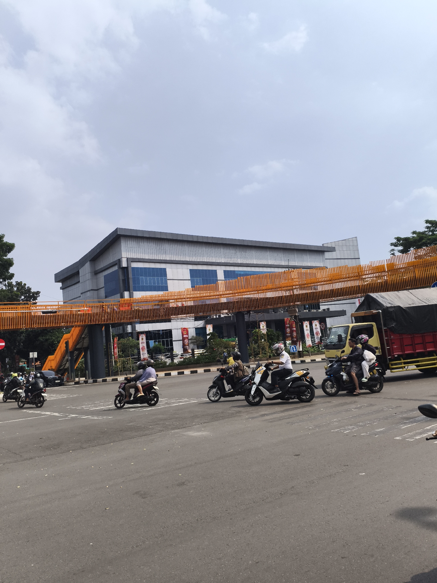 This image captures a daytime urban street scene in Cibinong, Indonesia, under a bright, overcast sky. The foreground shows an asphalt road with faded white markings, populated by numerous motorcyclists. Several scooters, varying in color (white, black, maroon), are visible, with most carrying one or two riders wearing helmets. One white scooter has "MASAGI" written on its side. The motorcycles appear to be at an intersection, possibly waiting for traffic to clear or starting to move.
In the midground, a yellow truck with a red flatbed and a dark tarp cover is positioned on the right side of the road. Spanning above the road is a prominent, bright orange pedestrian bridge with stairs leading up on the left, connecting towards a large, modern building in the background. This building features a sleek design with grey panels and prominent blue-tinted windows. Several banners hang near the building; one clearly displays "INDONESIA," another "SMART LIVING," and another partial text like "PERMATA." Green trees are visible on the far left and right edges of the frame, providing a natural contrast to the urban infrastructure. The scene depicts a typical busy day in a Southeast Asian city.