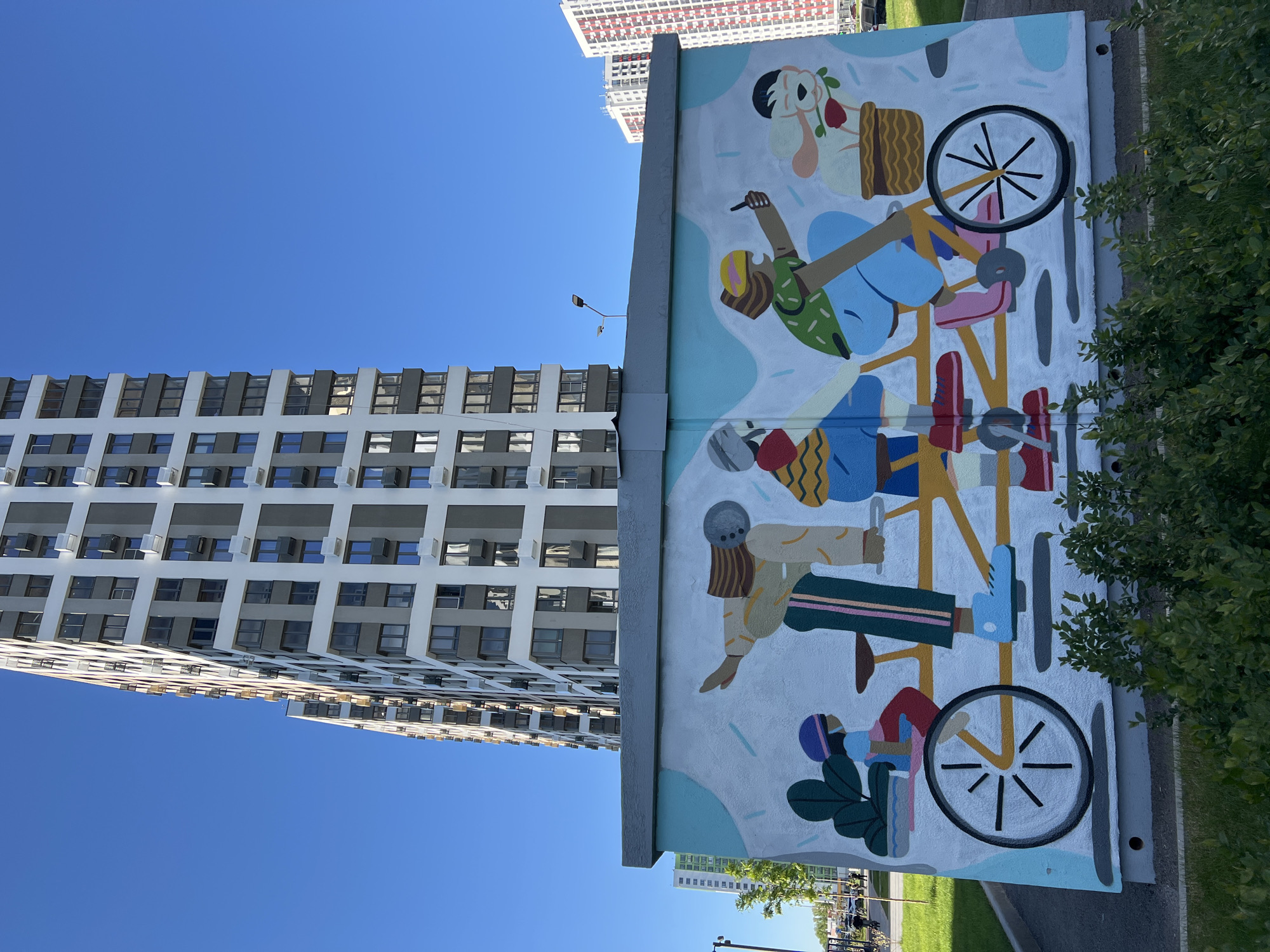The image depicts a mural on a wall in Ufa, Russia. The mural features an artistic representation of people riding a tandem bicycle. The style is cartoonish, with simplified shapes and bright colors. The figures are diverse and stylized.
Next to the mural is a tall residential building with many windows. The sky is clear and blue. Vegetation is visible in the foreground.