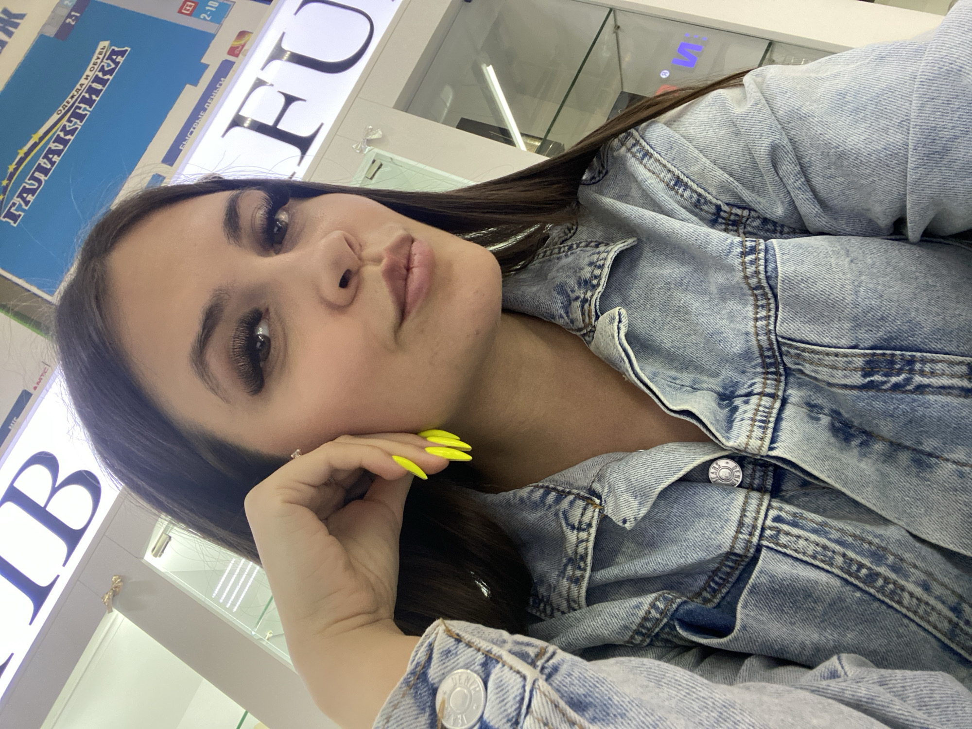 A medium close-up angled selfie shows a young woman inside what appears to be a shopping mall, based on the signs visible in the background. The woman has long dark hair, light skin, and wears long, false eyelashes. Her nails are painted a bright neon yellow. She's wearing a blue denim jacket. The background features a sign in Russian with Cyrillic characters, which appears to be “ТА ЛА ЛА ИКА” and advertises clothing and shoes. Another sign partially visible has “FU” on it. Given the location, this may be a store in the city of Chaikovsky, Russia. The woman is posing with her head resting on her hand. The overall lighting is bright and artificial, as is typical of indoor commercial spaces.