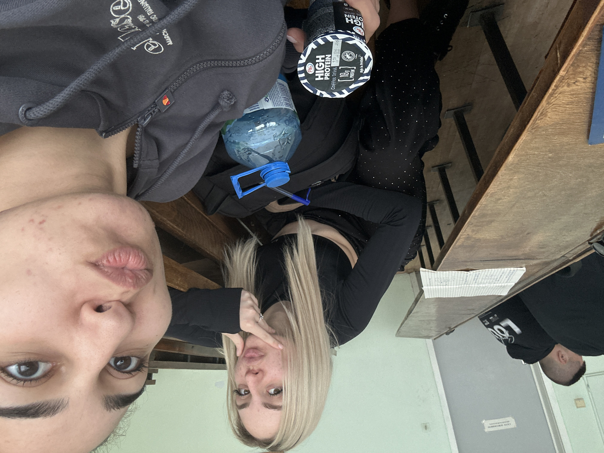 The image appears to be a selfie taken by one of two women sitting at a wooden desk, likely in a classroom setting. One woman is facing the camera directly, making a kissing face. She has dark hair, eyebrows, and eyes, and her skin has some blemishes. The other woman, with blonde hair and fair skin, is turned slightly and also making a kissing face while holding her hand near her face. Both women are wearing dark clothing.
On the desk, there is a gray hoodie, a bottle of water, and a container of "Pilos High Protein Coffee Drink." A pen is lying near the water bottle. The desks appear to be arranged in rows. To the right of the frame, a portion of another person is visible, wearing a black hoodie with a logo. The location in Burgas, Bulgaria, cannot be determined from the available image data.