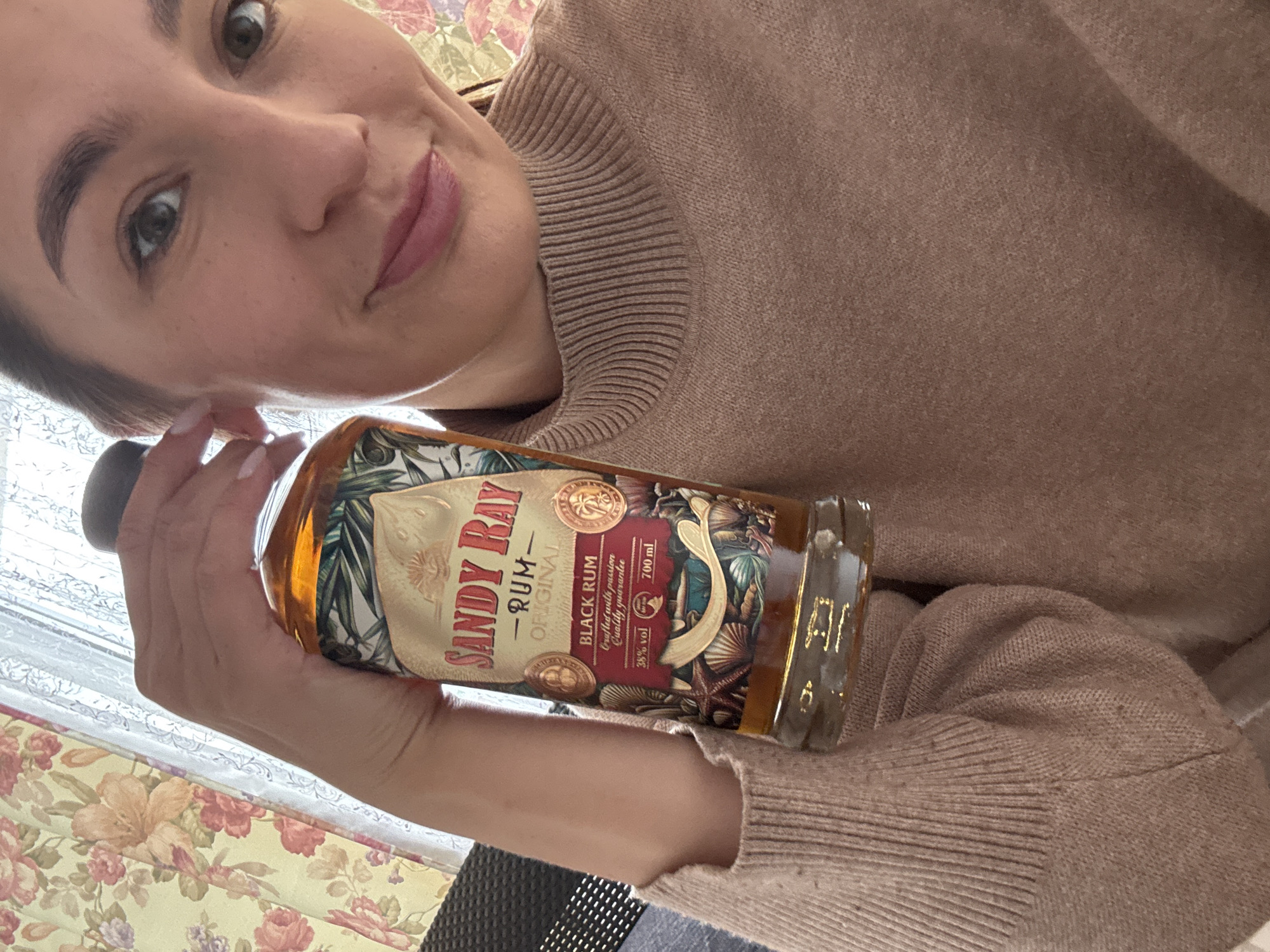 A woman is holding a bottle of "Sandy Ray" Black Rum, which is identified as being from Sochi, Russia based on the brand's origin. The bottle indicates it contains 700ml of rum at 38% alcohol by volume. The woman is wearing a tan sweater and is indoors, as indicated by the floral patterned window covering in the background. Her facial expression appears pleasant, with a slight smile.