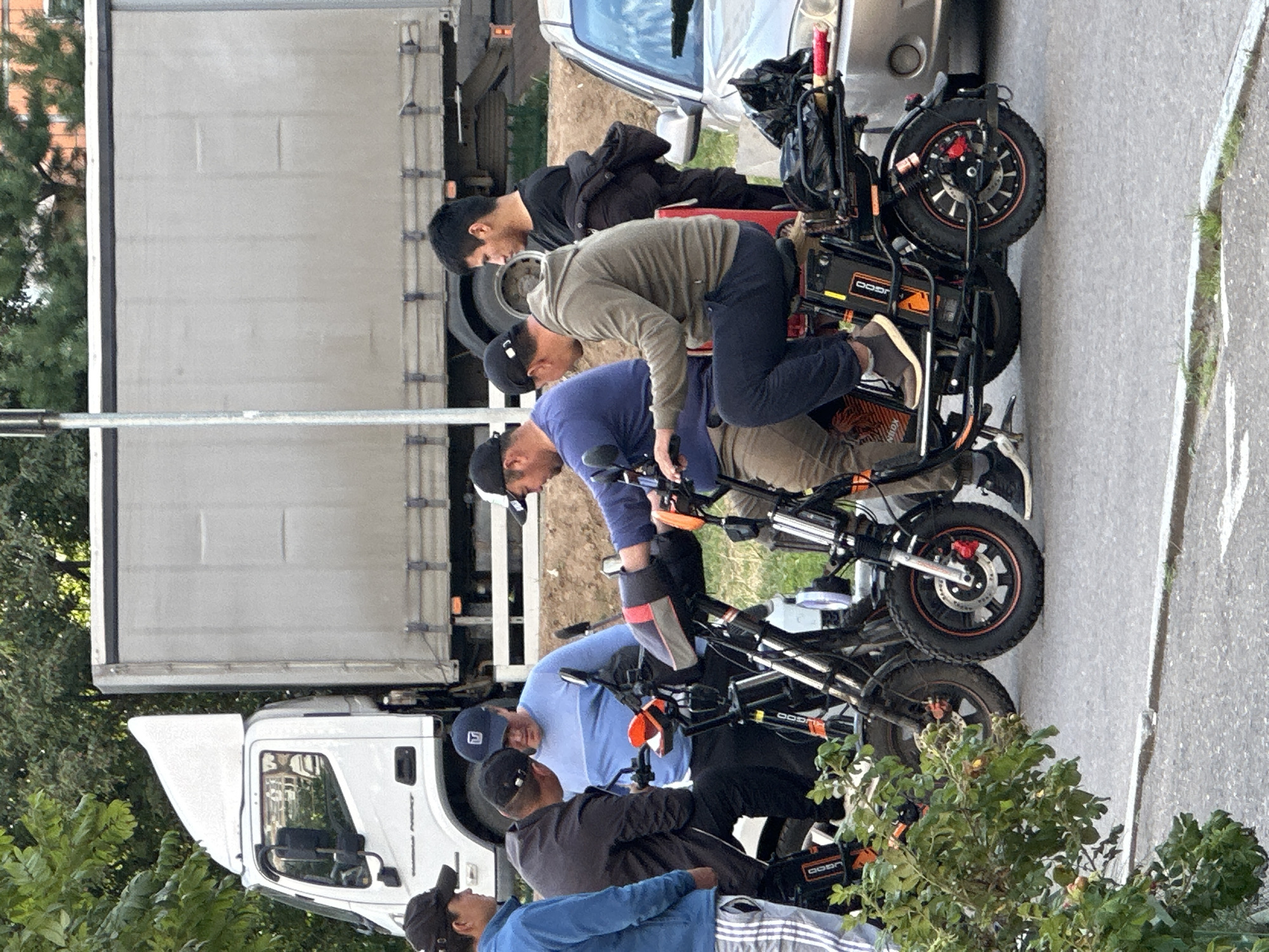 In Podolsk, Russia, a group of men are gathered around several electric scooters. A white truck with a gray cargo trailer is parked behind them, partially obscured by trees. To the right of the scooters, a silver car is visible. One man, wearing a blue jacket, stands on the left, while another in a black jacket stands next to him. Three men are seated on or near the scooters. One man is wearing a blue sweater and a baseball cap. Another, wearing a dark long-sleeved shirt and a baseball cap, is looking down at the scooter he is on. A third man, in a gray hoodie and dark pants, is seated on a scooter, looking forward. The environment suggests an outdoor setting, possibly a street or parking area.
