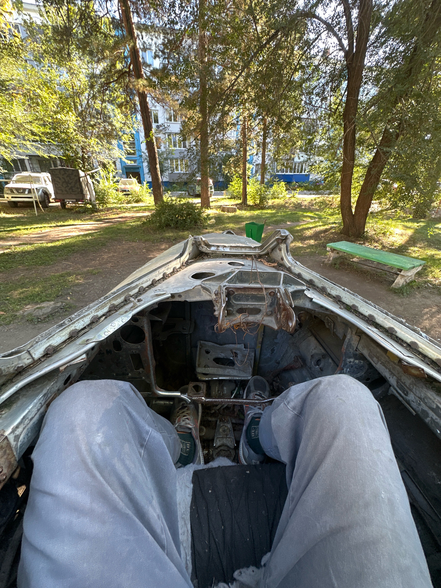The image depicts the interior of a derelict vehicle, viewed from the perspective of someone sitting inside. The car's interior is heavily damaged and appears to be a shell of its former self, with exposed metal, rust, and debris. The passenger's legs, clad in light grey pants, are visible, along with their feet wearing athletic shoes. In the background, a residential building with blue accents is visible through a canopy of trees. A green park bench and a green trash bin are also present in the outdoor setting. The scene suggests an urban environment, possibly a park or a residential courtyard in Biysk, Russia, given the context.