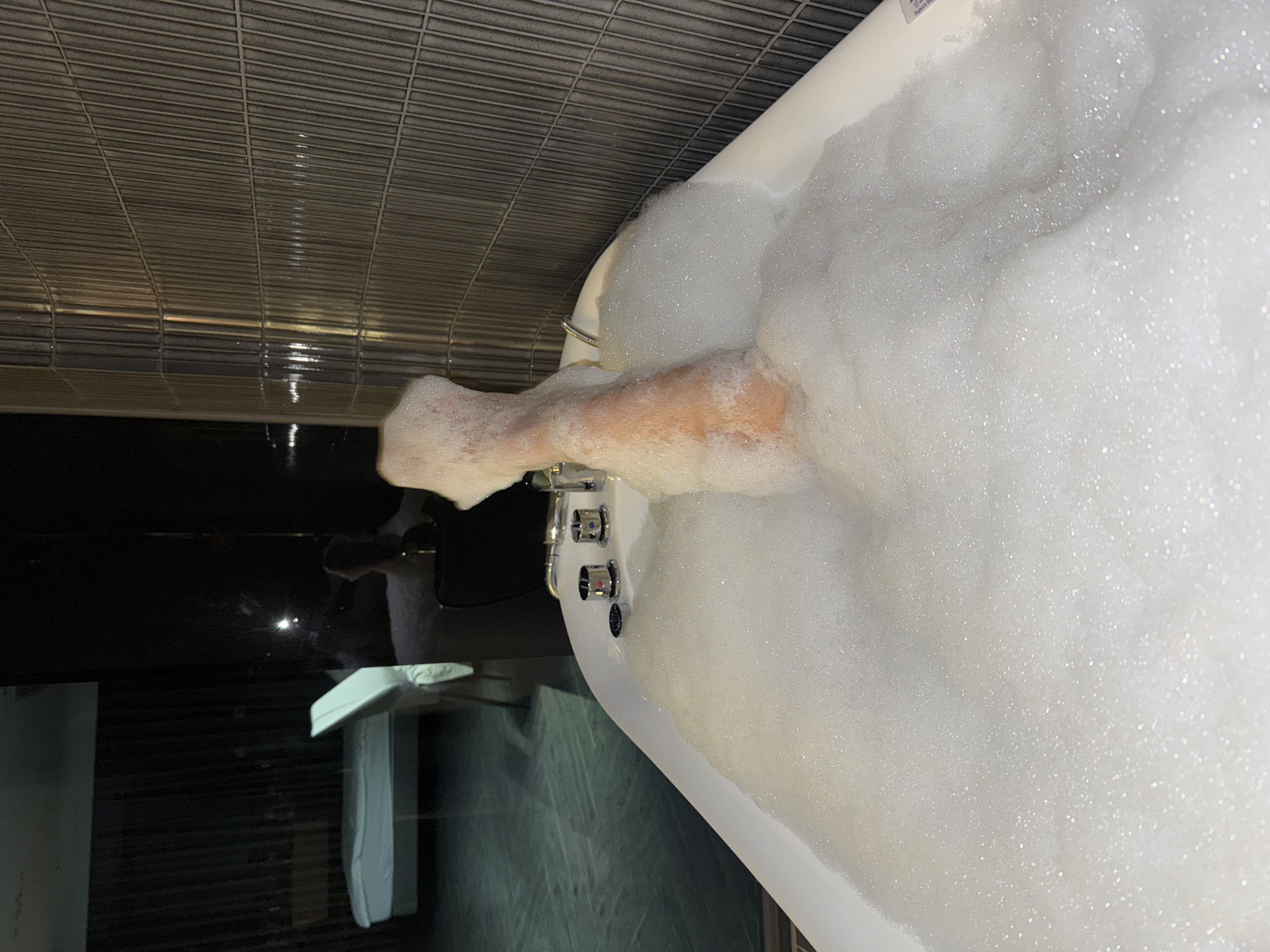 A partial view of a bathtub filled with white bubbles, obscuring most of the interior, is captured at an oblique angle. A human arm emerges from the bubbles, appearing somewhat submerged, with the hand positioned near the edge of the tub. The tub's faucet hardware is visible, featuring several control knobs. The surrounding environment includes a section of a wall covered in a grid-like texture, and a glimpse of what appears to be a cabinet or vanity unit with a white section. The city and specific location remain unknown.