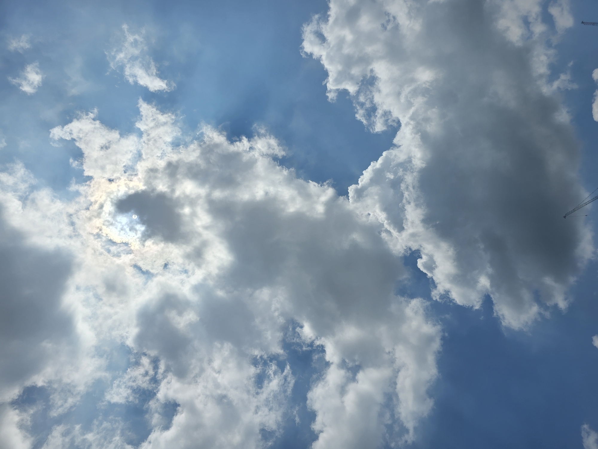 The image depicts a dramatic sky filled with a mix of bright white and darker gray clouds against a vibrant blue sky. Sunlight streams through a gap in the clouds, creating a luminous effect and visible sunbeams. In the lower portion of the image, two construction cranes are visible, suggesting an urban or industrial environment. The perspective is looking upwards. Based on the presence of construction cranes and the general visual aesthetic, this scene is consistent with a view from a developed area within Daejeon, South Korea, likely during daylight hours. There are no people or animals visible in the frame.