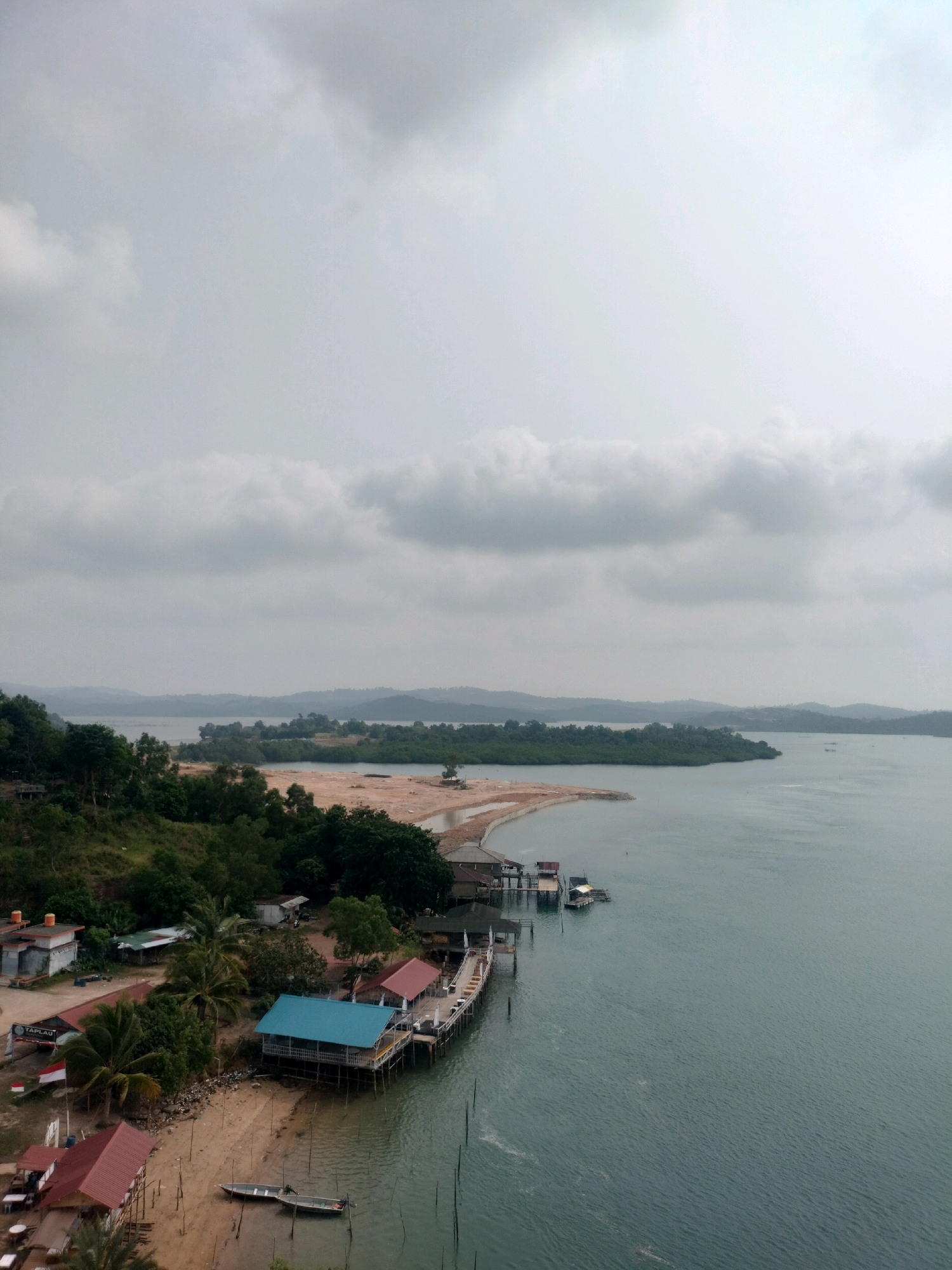 This aerial photograph captures a coastal scene in Batam, Indonesia, on an overcast day. The image is dominated by a wide expanse of calm, gray-green water on the right, meeting a sandy shoreline dotted with buildings and vegetation on the left.
The shoreline itself is varied. In the foreground on the left, a cluster of buildings with red and blue roofs are situated on a sandy beach. One building, with a bright blue roof, extends out over the water on stilts, forming a pier or boardwalk. Another building with a red roof also appears to be over the water, supported by wooden stilts. Several small boats, including a metallic silver one with an outboard motor, are beached or moored near the shore. Further down the beach, more buildings are visible, some appearing to be residences and others possibly commercial structures. A red and white flag, likely the Indonesian flag, flies near one of the buildings. Palm trees and other lush green vegetation are interspersed among the structures.
Beyond this immediate coastal area, the land rises into a densely forested hillside on the far left, with more distant hills forming the horizon. The water extends to the middle ground, where more islands with green vegetation are visible, some appearing as strips of land. A few small, dark shapes, possibly fishing platforms or distant boats, are scattered on the water in the middle distance.
The sky is filled with heavy, gray clouds, contributing to the muted light and somber atmosphere of the scene. The overall impression is one of a tropical, coastal settlement, likely a village or a small town, situated along a tranquil body of water. The presence of stilted structures over the water suggests a location that experiences tides or is built to accommodate the proximity of the sea.