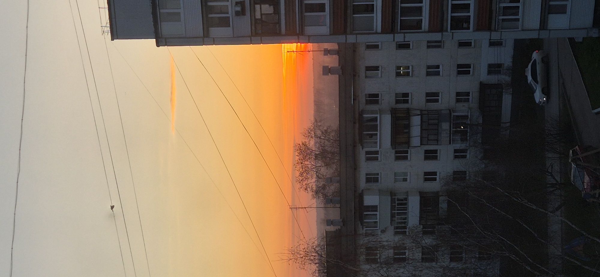 The image captures a vibrant sunset or sunrise over an urban or suburban landscape. The sky dominates the top and central portions, displaying a striking gradient of colors from pale yellow-orange on the top left to intense orange and deep red nearer the center-right. Several dark, thin power lines or utility cables stretch horizontally across the sky, with a small, dark, heart-shaped object notably hanging from one of them. A faint, elongated, lighter streak, possibly a contrail, is visible in the upper left sky.
To the right, two multi-story residential buildings stand, partially silhouetted against the colorful sky. The building in the foreground has a darker facade, while the larger building behind it is lighter-toned with numerous rectangular windows and balconies, some with enclosed glass. Bare, leafless trees are scattered between and in front of the buildings, their branches forming intricate dark patterns.
At the bottom right, a white car is parked on what appears to be a street or parking area, its taillights visible, suggesting the low light conditions of dusk or dawn. Faint structures, possibly part of a playground, are discernible in the far bottom right. The scene is devoid of people or direct activity, presenting a still and atmospheric view of natural beauty in an urban setting during either dawn or dusk. There is no visible text in the image.
