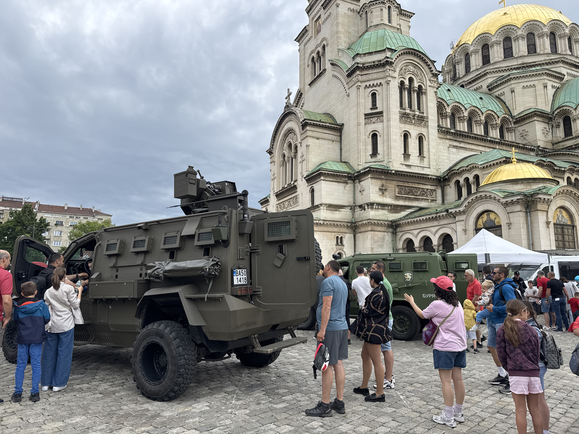 The image depicts a bustling scene in front of a large, ornate building, likely a cathedral, identifiable by its distinctive architecture and golden domes. The scene is set on a paved plaza with a cloudy sky overhead.
The focal point of the image is a large, olive-green armored military vehicle. People are gathered around the vehicle, with some individuals interacting with it, either looking inside or taking pictures.
In the background, there's another similar, though smaller, armored vehicle. There is also a white tent nearby.
The location is in the city of Sofia, Bulgaria, as indicated by the Cyrillic writing (БУРЯ) on the side of the vehicle and the plate number BA50.