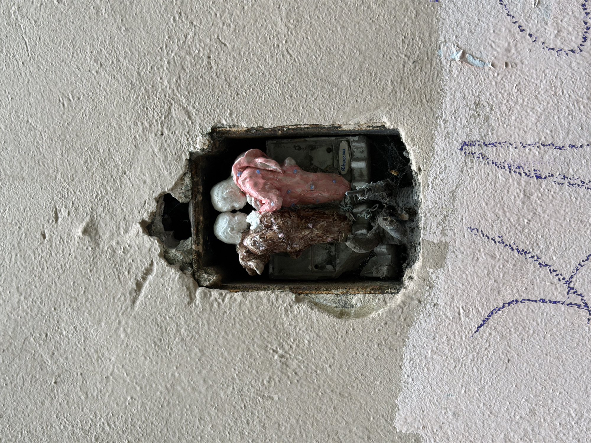 An eye-level shot reveals a rectangular cavity in a rough, painted wall, likely plaster or stucco. The hole exposes the interior of a defunct electrical box. Inside the box are remnants of electrical components, including a fuse box, wires, and what appear to be globs of colorful sealant or clay. The walls are damaged and flaking with blue graffiti. This structure is in Saint Petersburg, Russia.