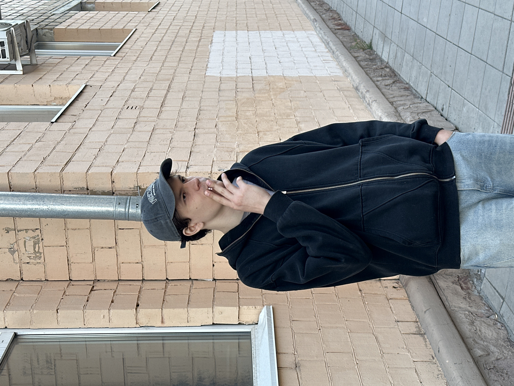 A young person wearing a gray baseball cap, black zip-up hoodie, light blue jeans, and a white t-shirt is standing outside and appears to be smoking. Their right hand is near their mouth, holding an object. The location appears to be an urban setting with brick buildings and a sidewalk. A metal pipe runs vertically along the wall. Based on architectural styles and common city layouts, the location could be in Krasnoyarsk, Russia. The person is standing on a paved surface next to a building made of light-colored bricks. The building has windows with metal frames. The sidewalk is adjacent to the building and is made of square paving stones. There is also a concrete curb.