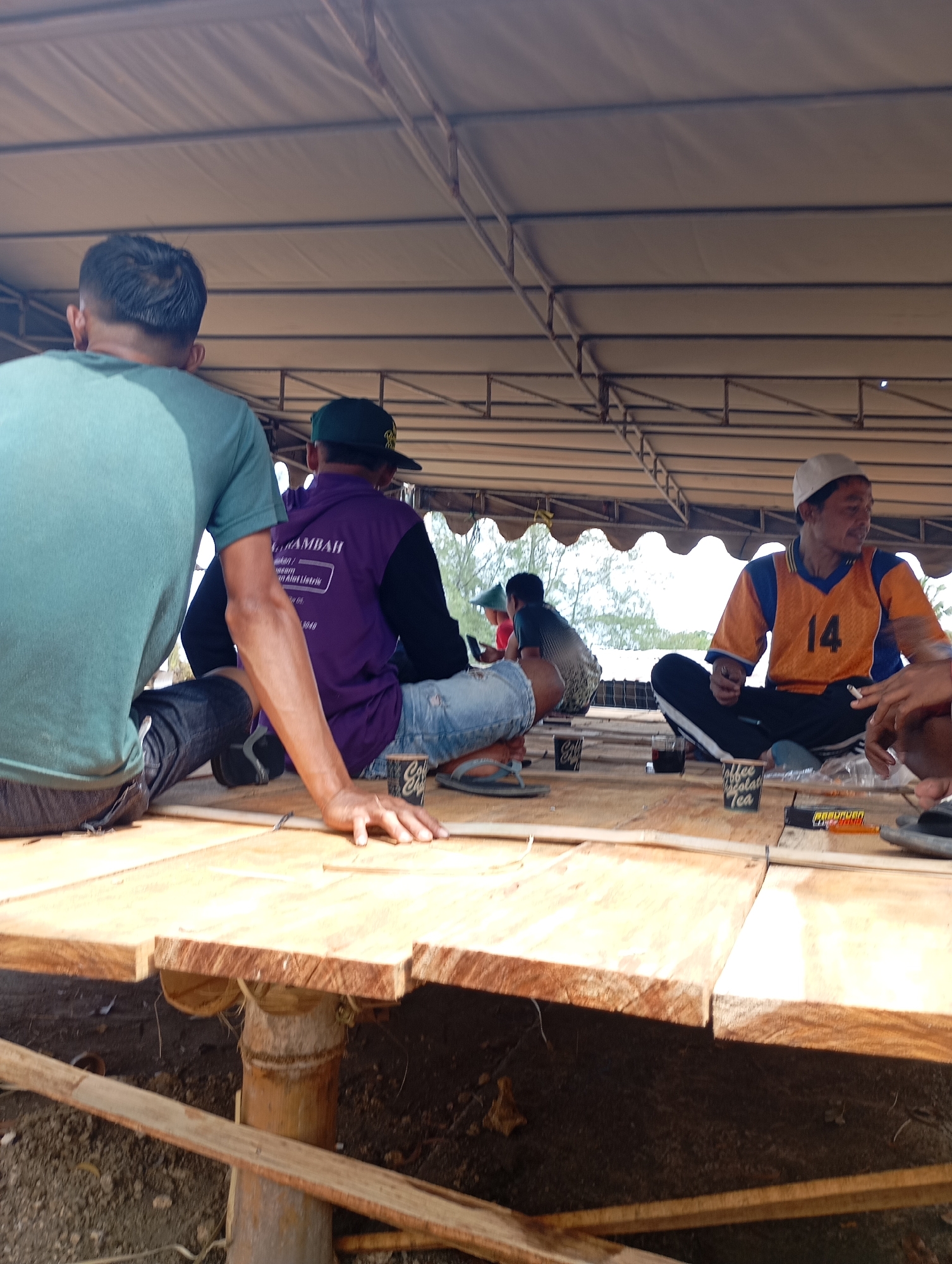 In the city of Ketepung, Indonesia, under a large tent structure supported by metal beams, several individuals are seated on a raised wooden platform. The platform appears to be constructed from rough-hewn wooden planks. In the foreground, a person wearing a teal t-shirt and dark jeans is seated with their back mostly to the camera, their arm resting on the platform. To their right, another individual is wearing a purple long-sleeved shirt with text on the back and denim shorts. Further to the right, a man in an orange and blue jersey with the number "14" and a white skullcap is seated. He is holding a cigarette. Between the individuals, there are at least three small paper cups on the platform, some containing dark liquid, and a pack of cigarettes. In the background, other individuals are visible, and trees and foliage can be seen through the gaps in the tent. The overall scene depicts a casual gathering or rest stop.