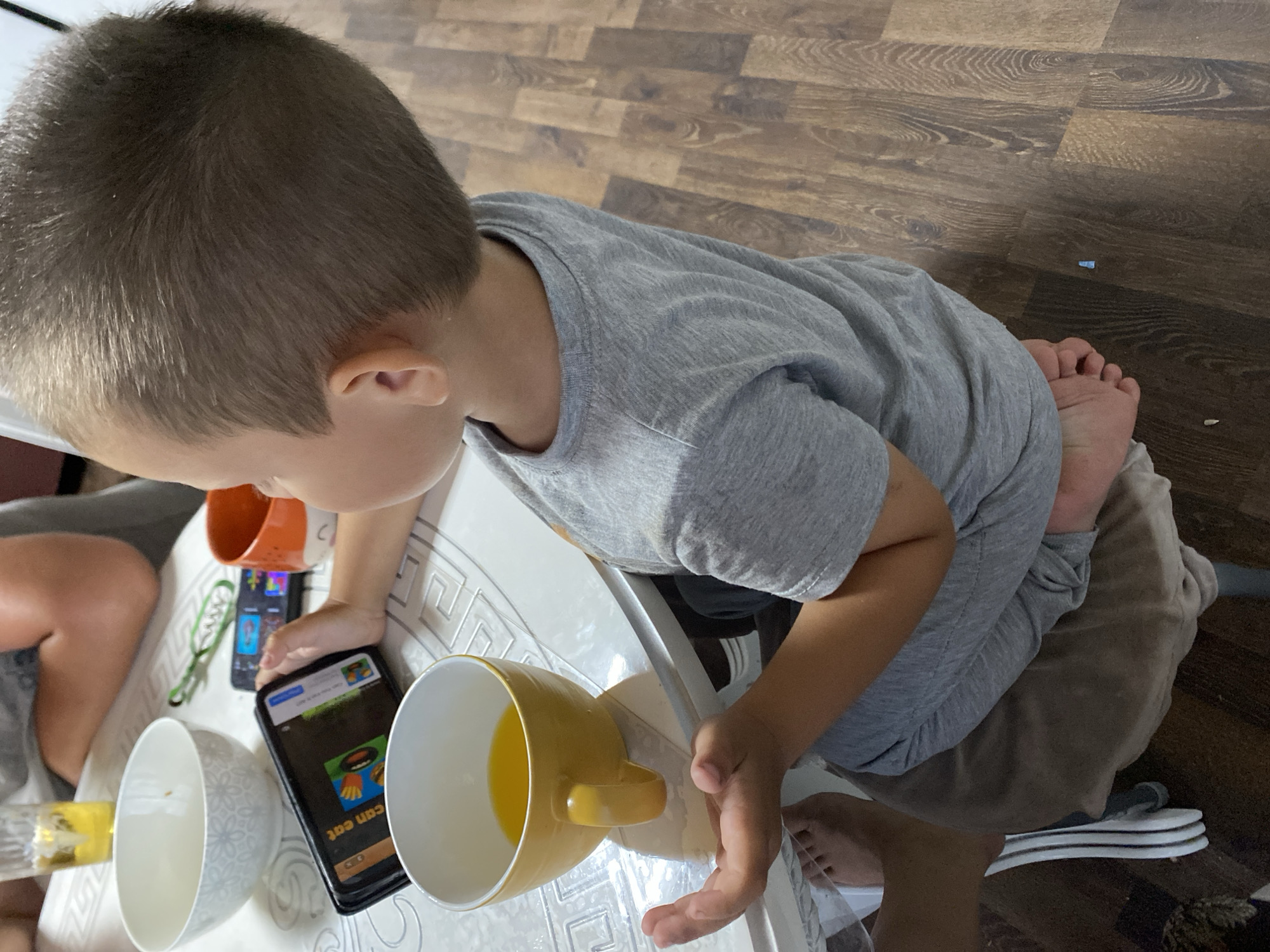 A young boy is seated at a table, engrossed in playing a game on a smartphone. He is wearing a grey t-shirt and grey sweatpants. His left hand rests on the phone, while his right hand is open, palm up. In front of him is a yellow mug filled with orange juice, and next to it, a white bowl. Another smartphone and an orange mug are visible further down the table. The table itself has a patterned surface. The boy's feet are visible at the bottom of the frame, resting on a brown cushion. The setting appears to be a dining area, with a wooden floor. The image provides no information to suggest its location in Chirsha-Tartysh, Russia.